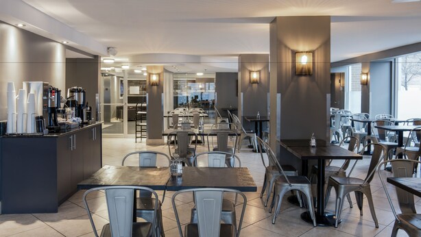 A modern dining area with multiple tables and chairs, a coffee station, and large windows providing natural light.