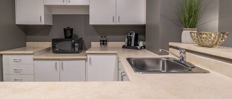 Una cocina moderna con armarios blancos, un lavamanos de acero inoxidable, microondas, cafetera y una planta en maceta.