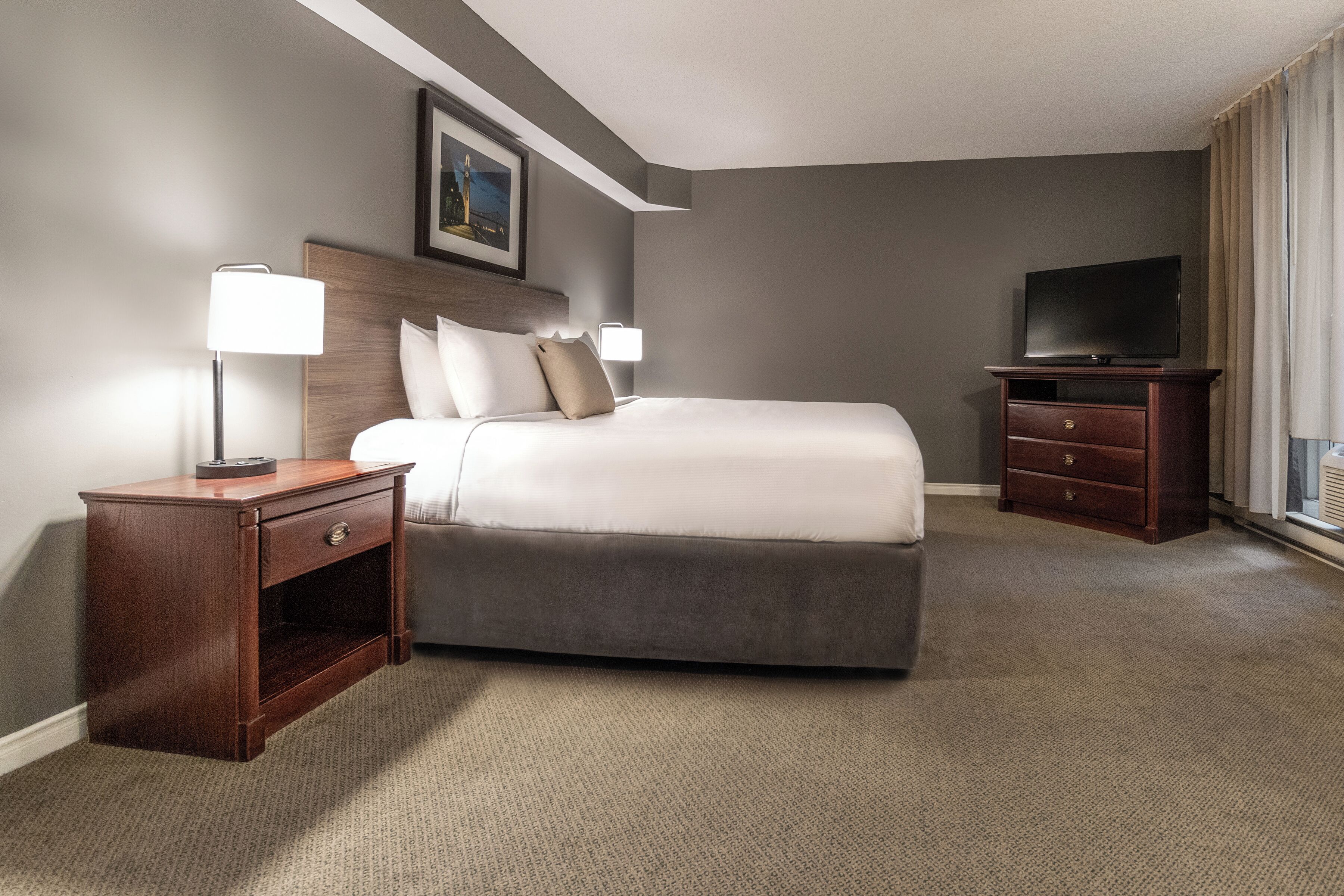 A hotel room with a bed, a nightstand, a TV, and a painting on the wall.