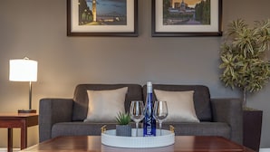 A modern living room with a sofa, a coffee table with a bottle and glasses, framed pictures on the wall, and a lamp.