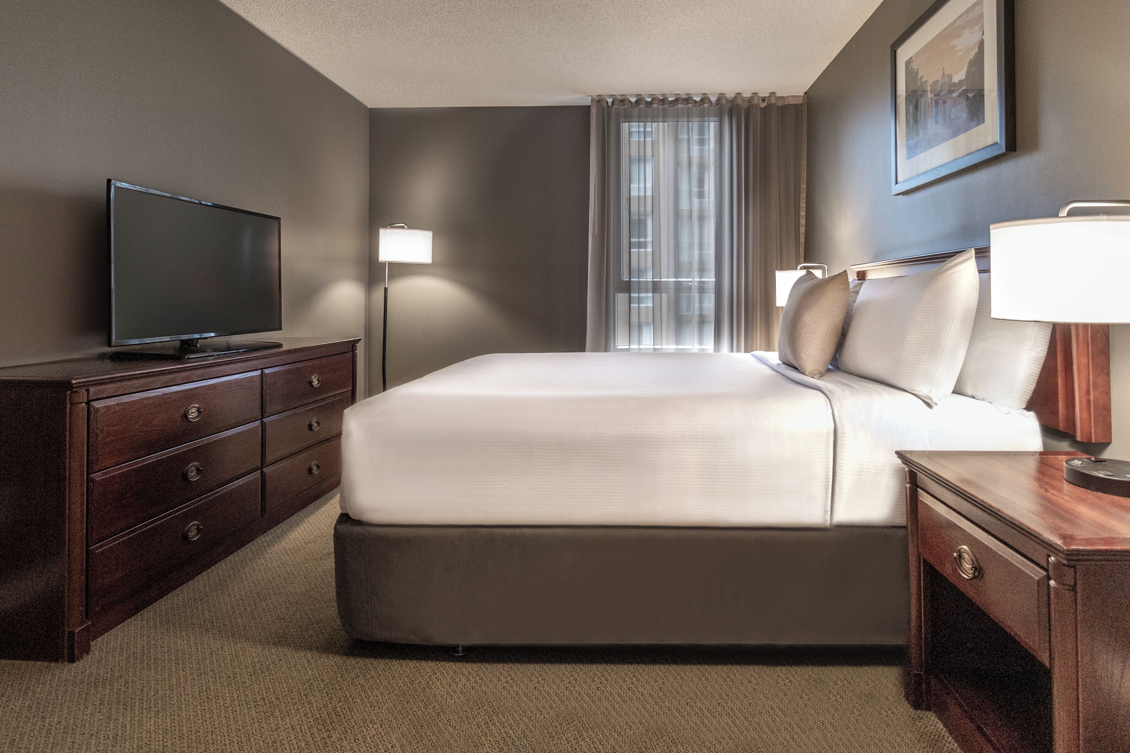 Une chambre d’hôtel avec un grand lit, un téléviseur à écran plat posé sur une commode en bois, une table de chevet avec une lampe et une fenêtre avec des rideaux.