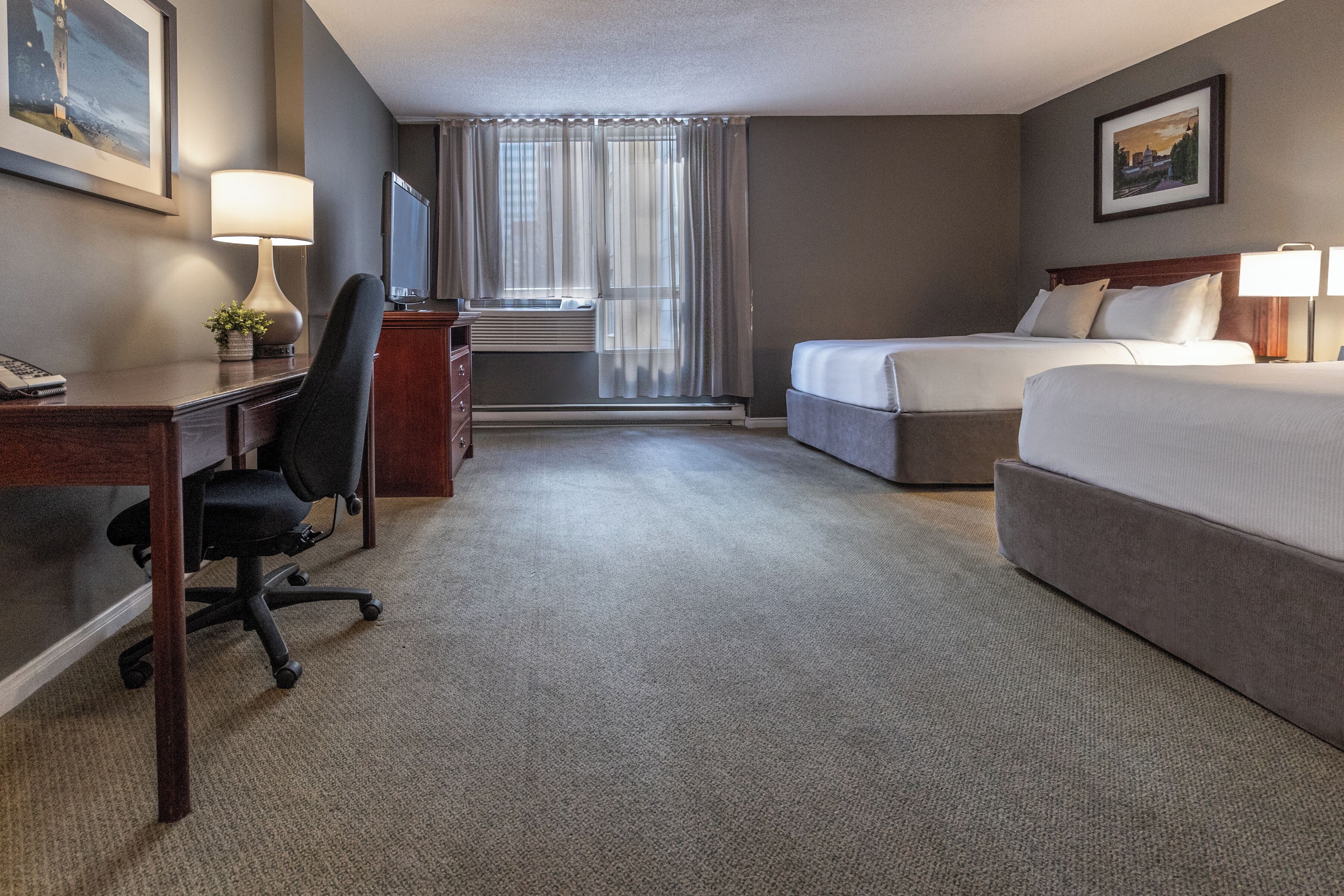 Quarto de hotel com escrivaninha, duas camas, uma cadeira, luminária, televisão e janela com cortinas.