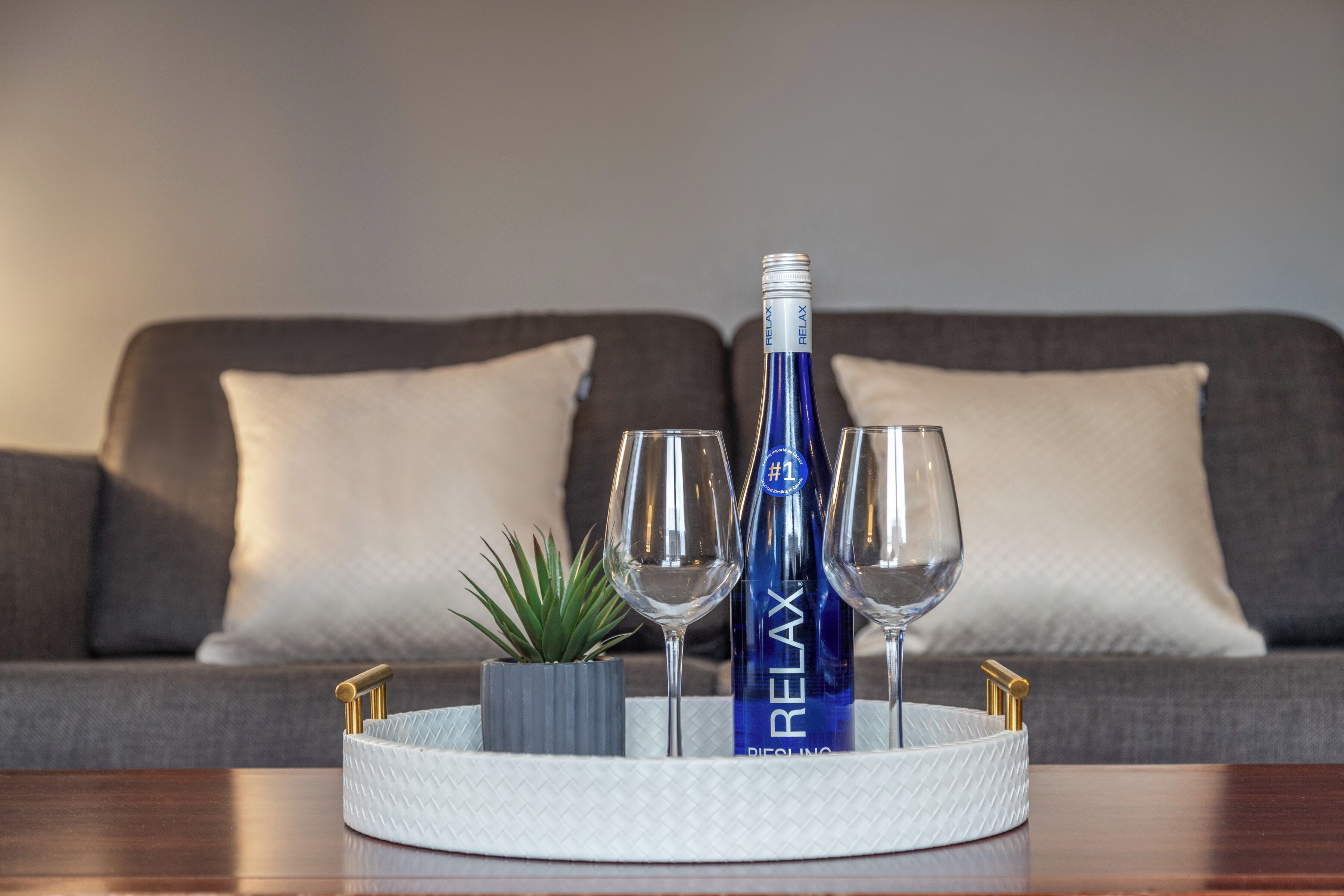 A tray with two wine glasses, a bottle labeled "RELAX," and a small plant on a wooden table in front of a grey sofa with white cushions.