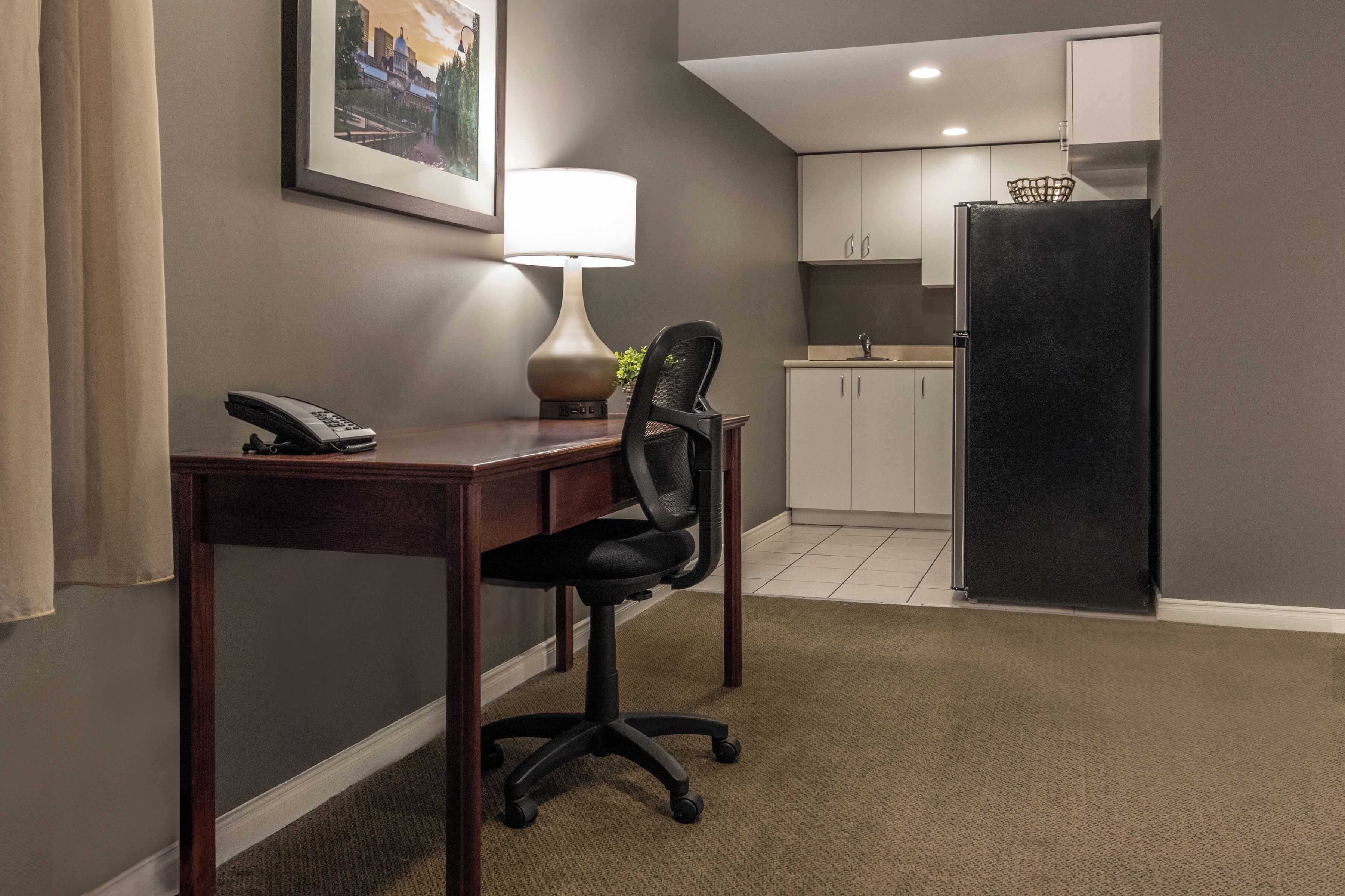 A hotel room with a desk, chair, lamp, phone, and a kitchenette with a refrigerator.