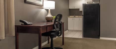 A hotel room with a desk, chair, lamp, phone, and a kitchenette with a refrigerator.