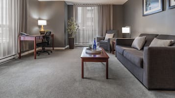 Una habitación de hotel moderna con un sofá gris, una mesa de centro de madera, un escritorio con una lámpara, una planta en maceta y un gran ventanal con cortinas.
