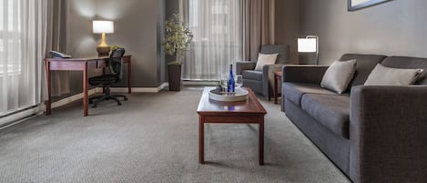 Una habitación de hotel moderna con un sofá gris, una mesa de centro de madera, un escritorio con una lámpara, una planta en maceta y un gran ventanal con cortinas.