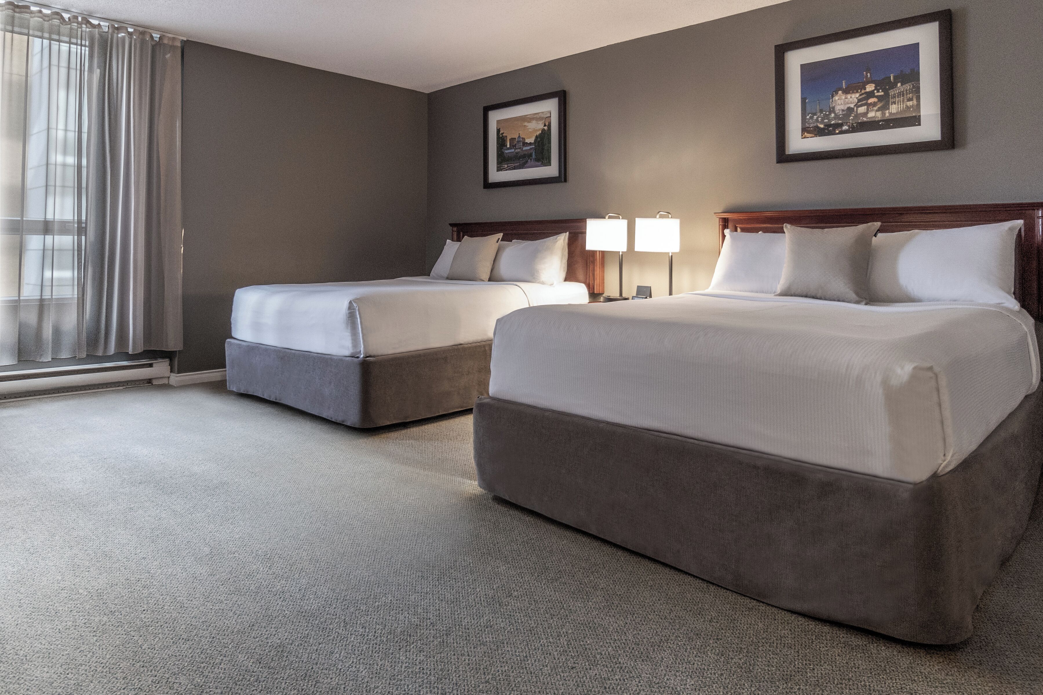 A hotel room with two beds, a large window with curtains, a framed picture on the wall, and a carpeted floor.