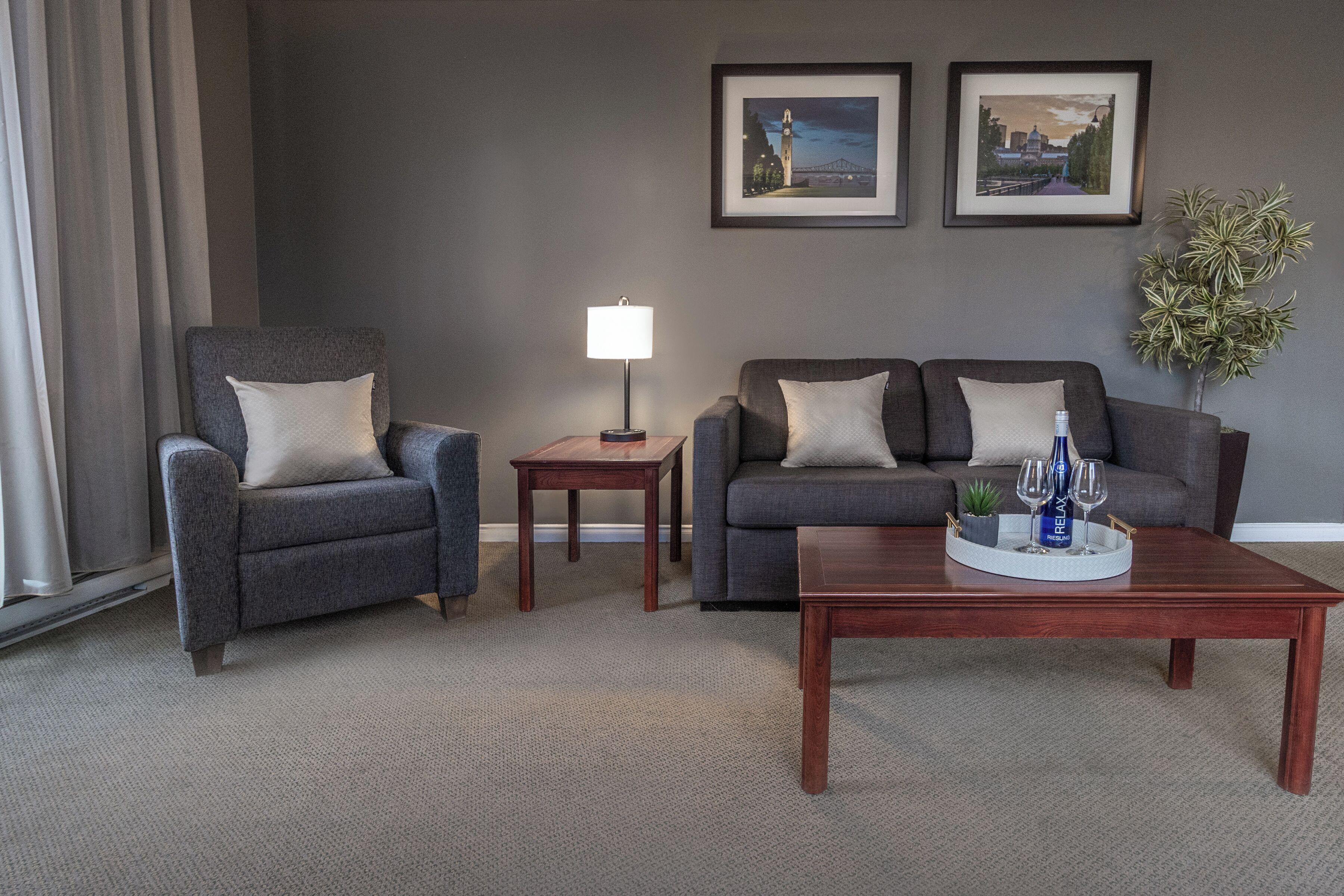 A modern living room with a grey sofa, a matching armchair, a wooden coffee table with a tray of glasses and a bottle, a small side table with a lamp, and two framed pictures on the wall.