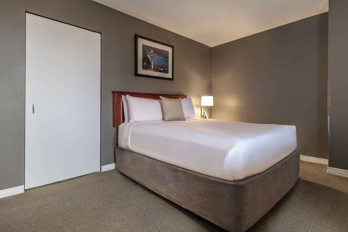 A hotel room with a large bed, a door, a framed picture on the wall, and a nightstand with a lamp.