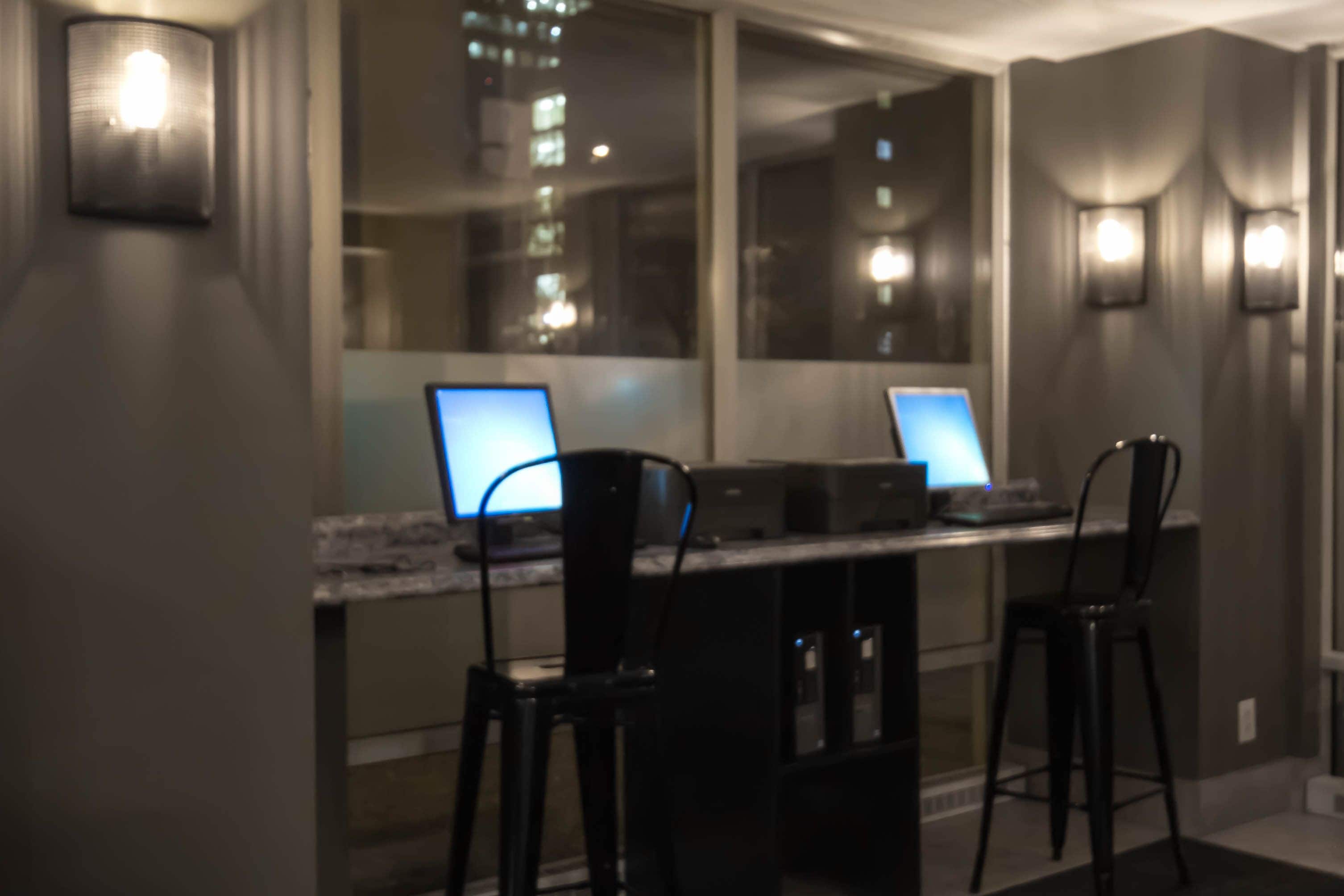 A modern kitchen with two computers on a counter, black bar stools, and a large window with city lights visible outside.
