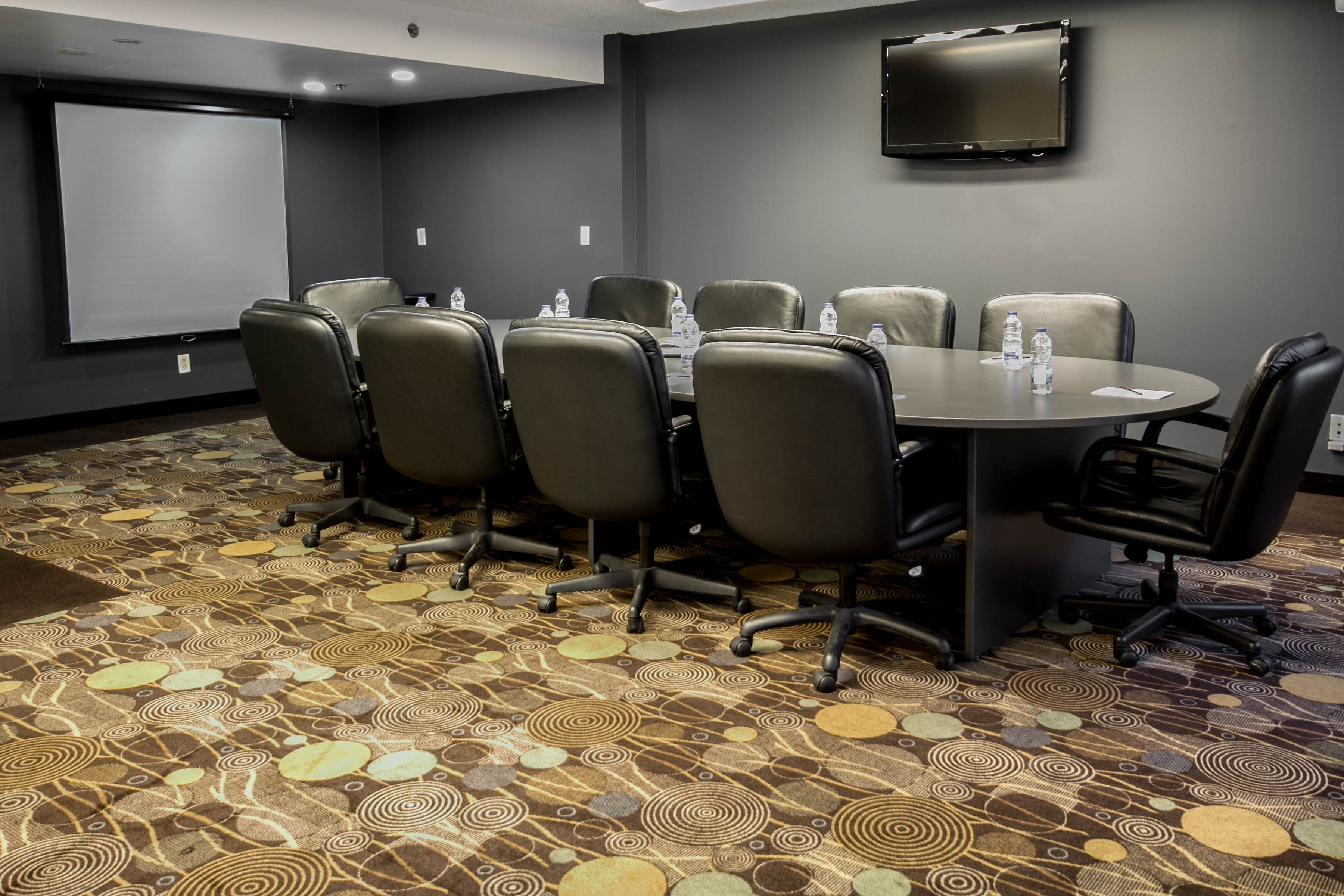 A conference room with a round table, six chairs, a projector screen, and a flat-screen TV on the wall.