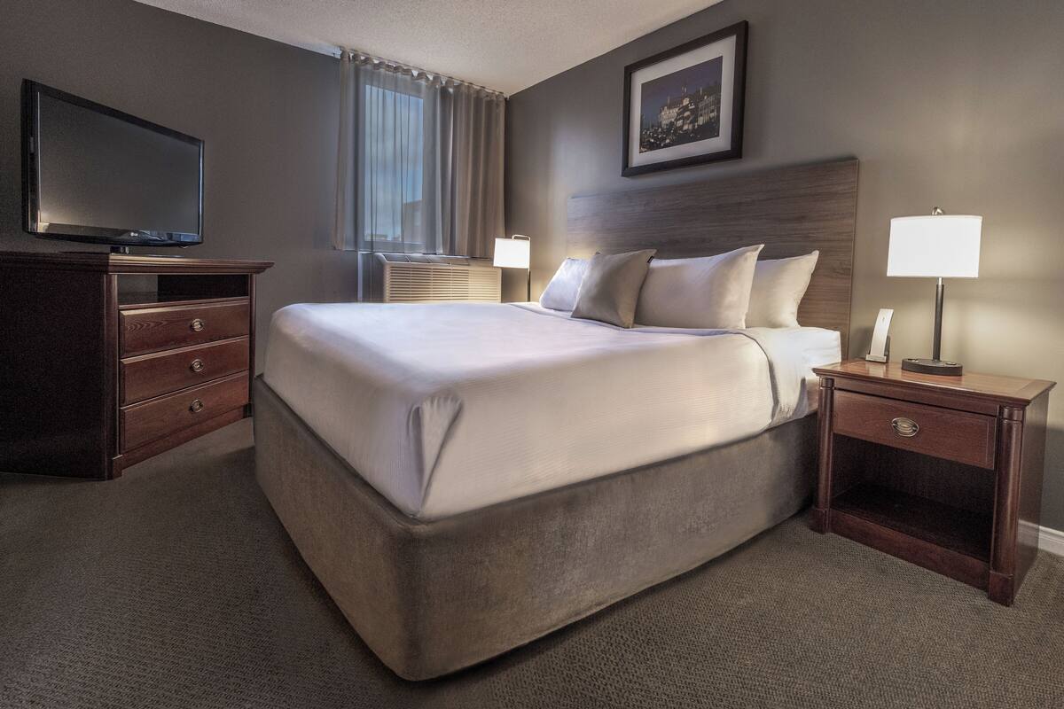 A hotel room with a large bed, a TV on a dresser, a nightstand with a lamp, and a window with curtains.