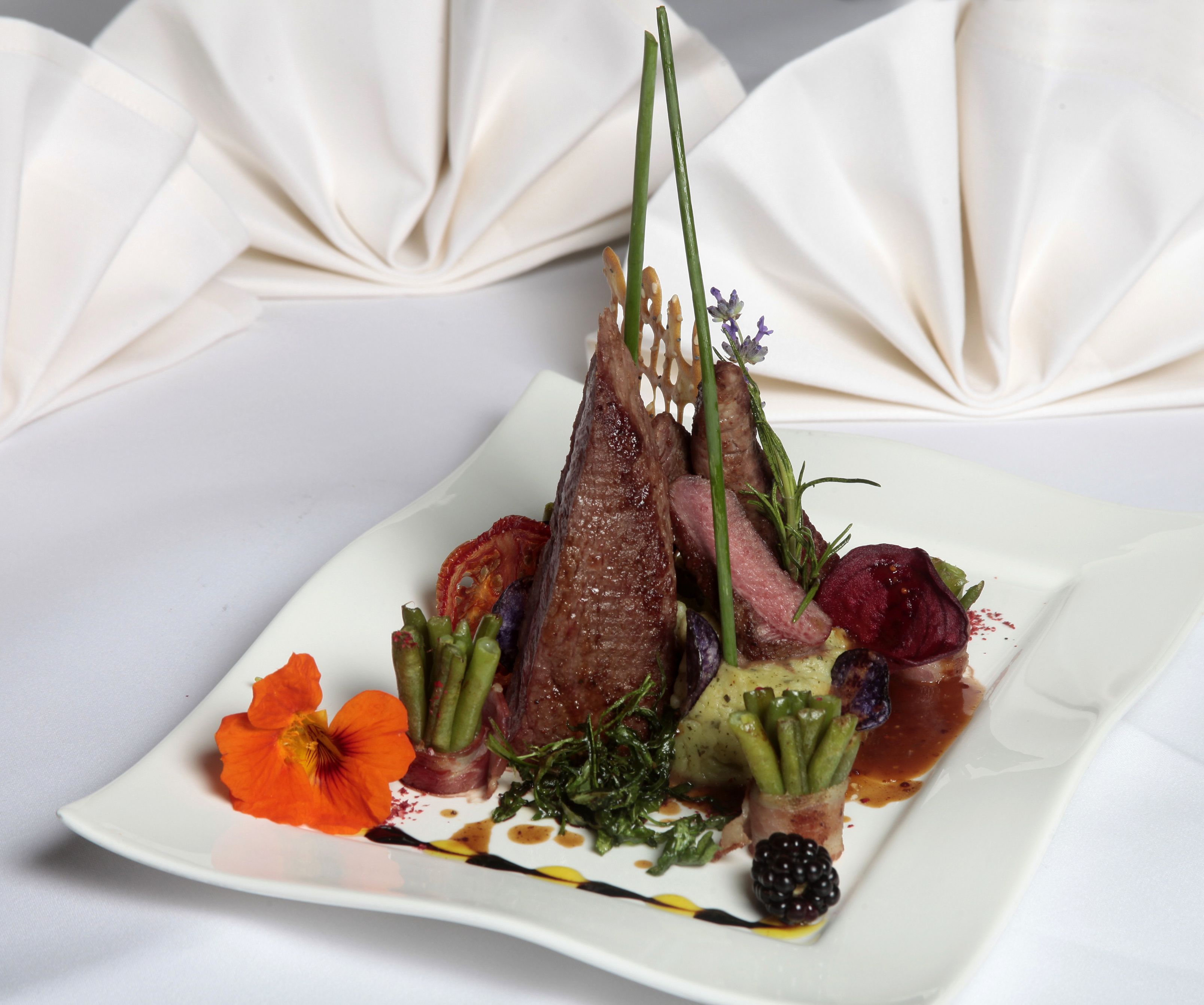A square white plate with a variety of food items including asparagus, a piece of meat, and a flower.