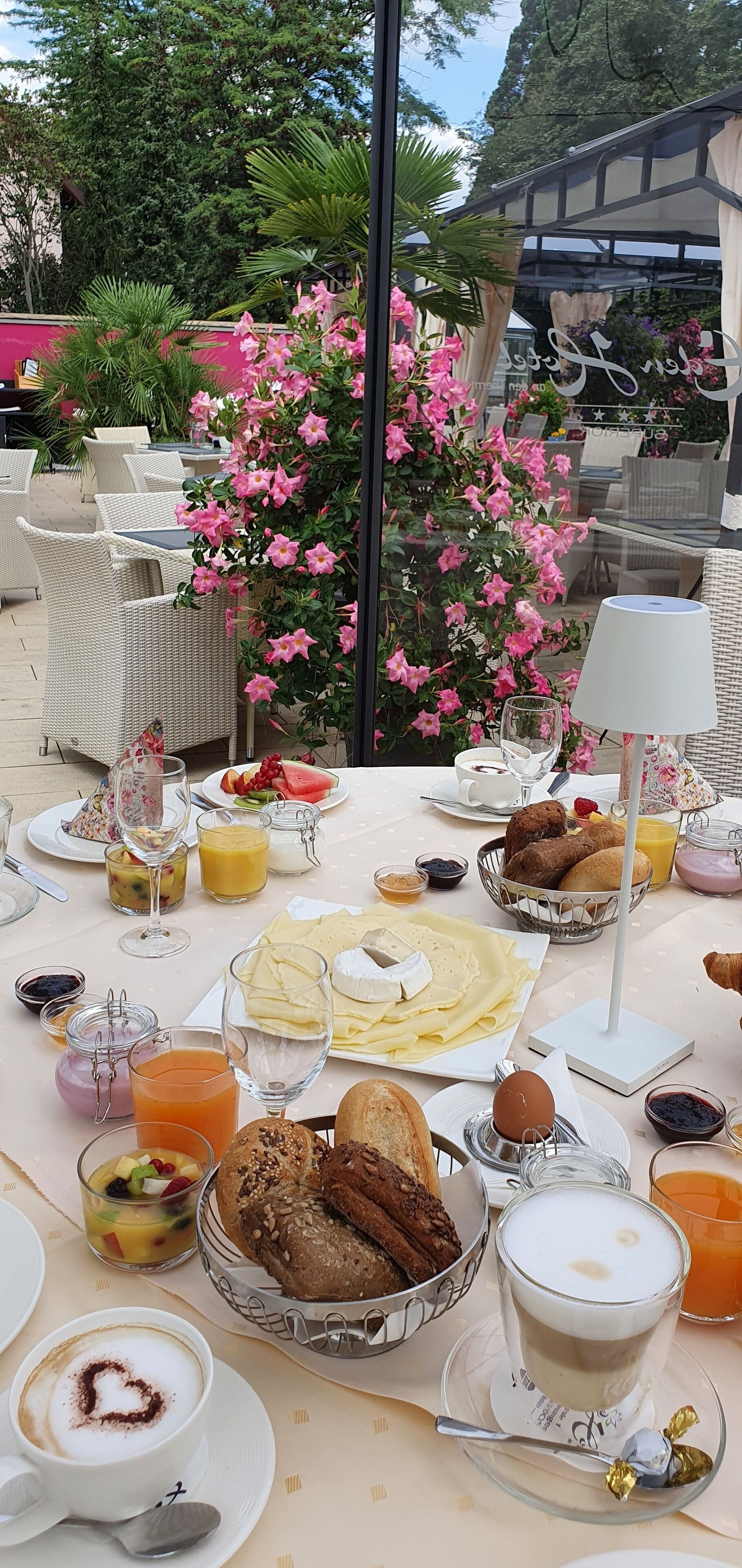 An outdoor dining area with wicker furniture, a variety of breakfast items, and a large pink flowering plant.