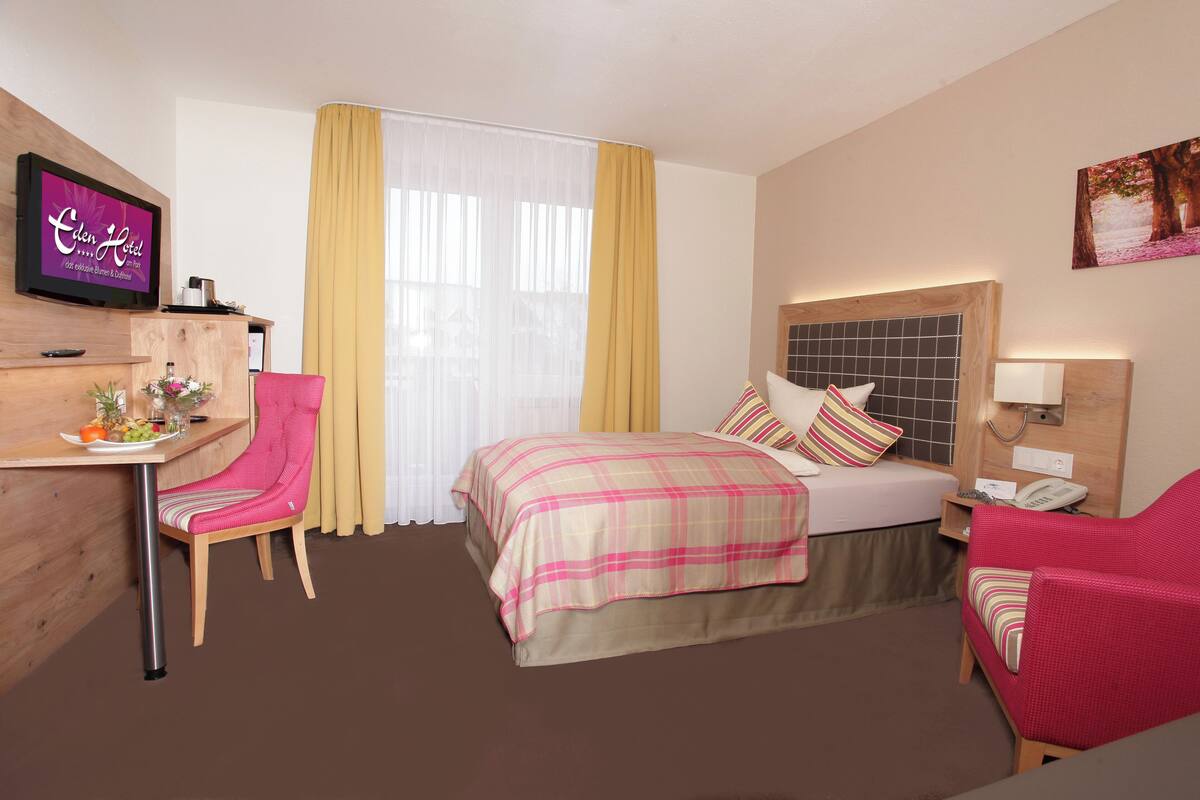 Quarto de hotel com cama, escrivaninha, cadeira, televisão e um quadro na parede.