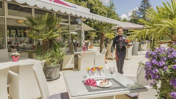 An outdoor dining area with wicker furniture, potted plants, and a waiter carrying a tray.