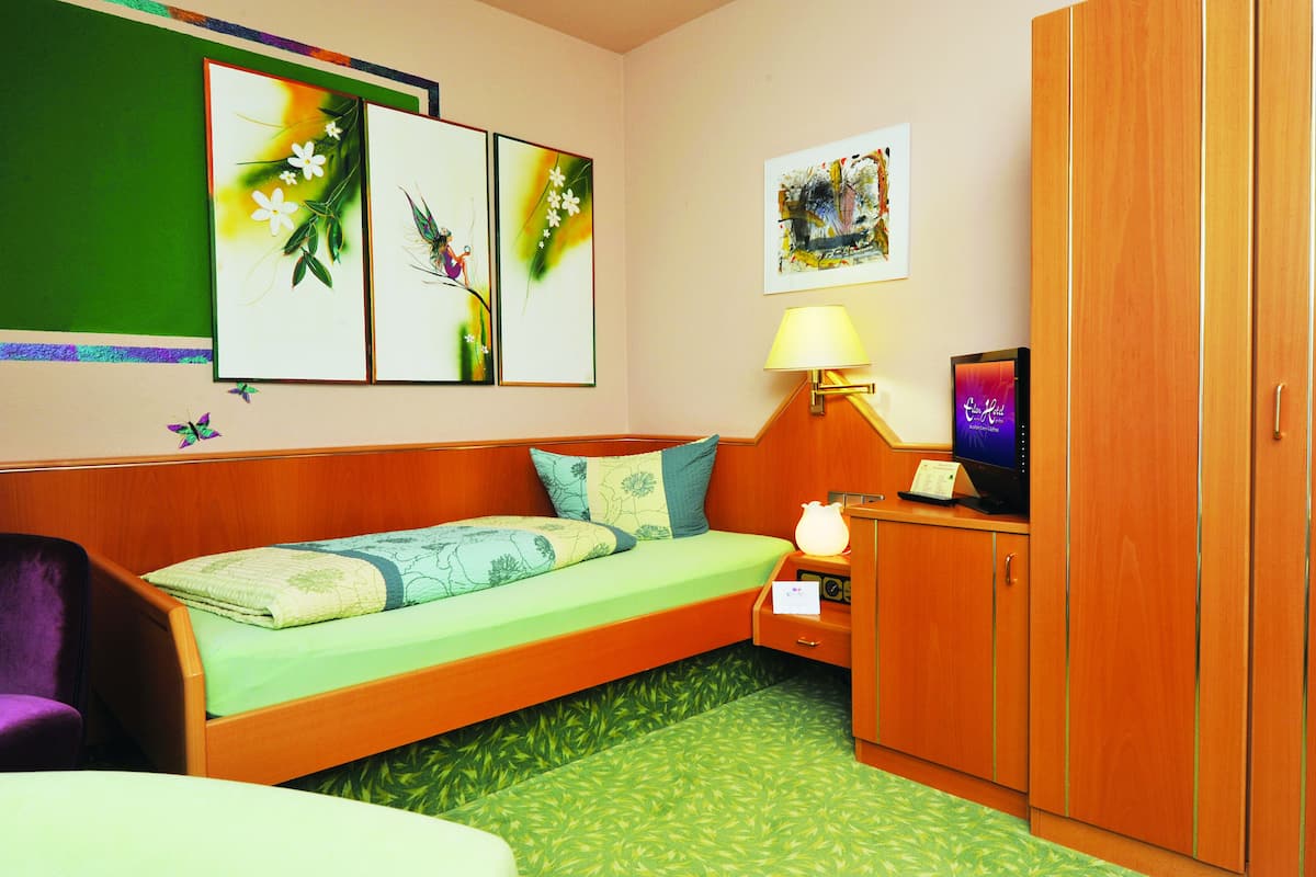 Quarto de hotel com cama, escrivaninha iluminada por uma luminária, televisão e obras de arte nas paredes.