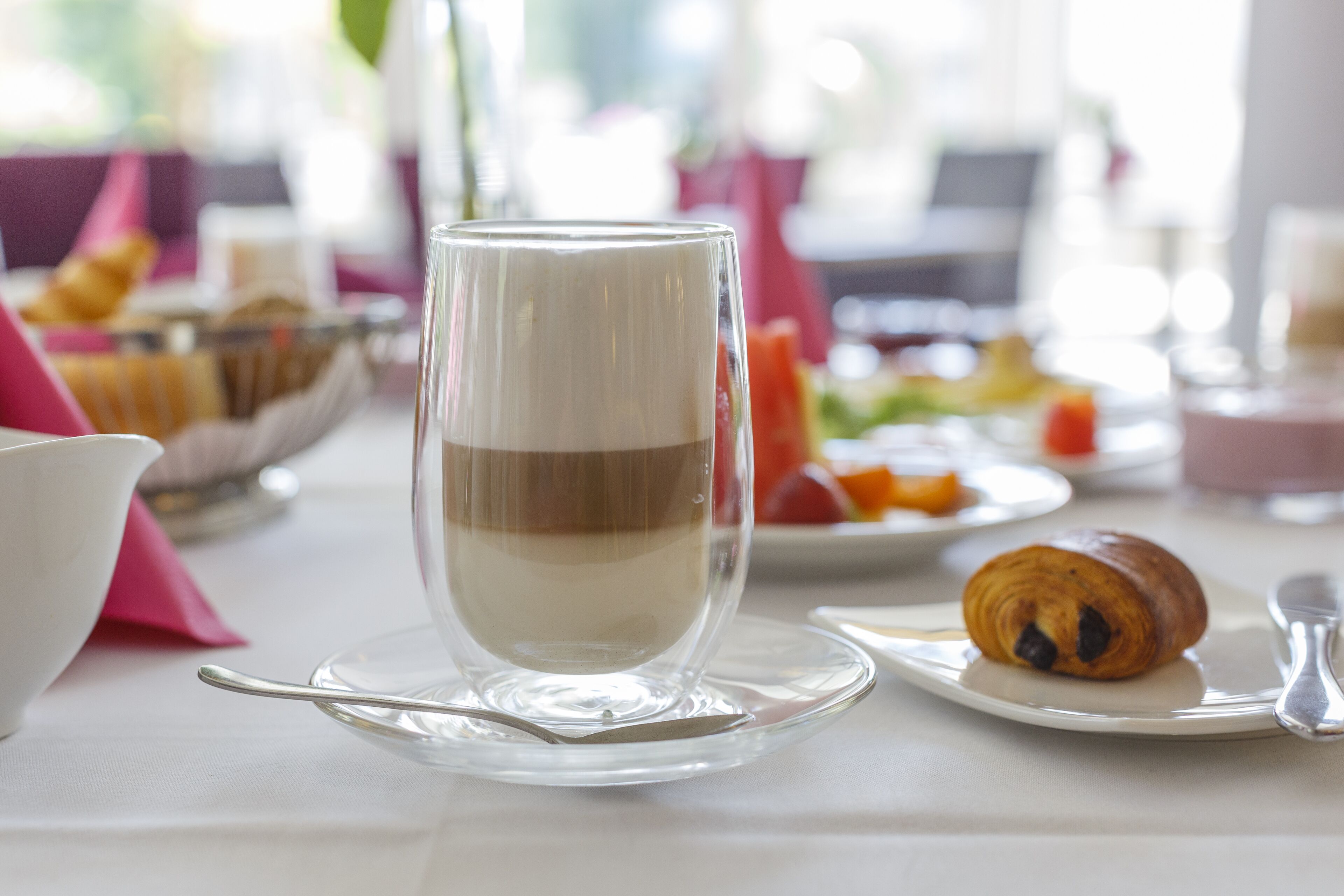 Una bebida en capas en un vaso transparente sobre un platito, un croissant, y un plato con fruta sobre una mesa.