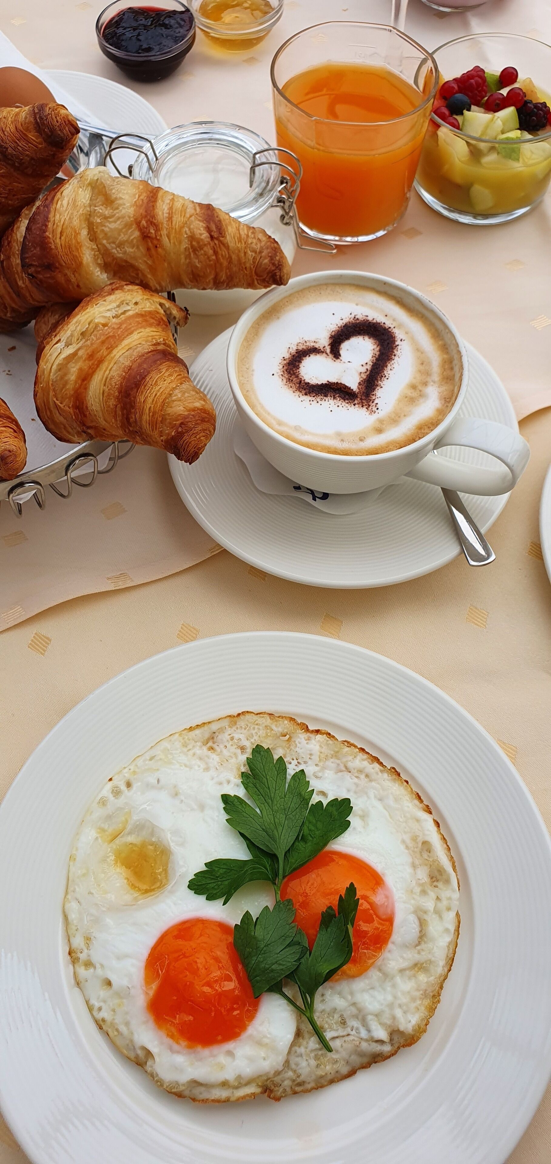 A breakfast setting with croissants, a cup of coffee with heart-shaped latte art, a glass of orange juice, and a plate with a fried egg and tomato.