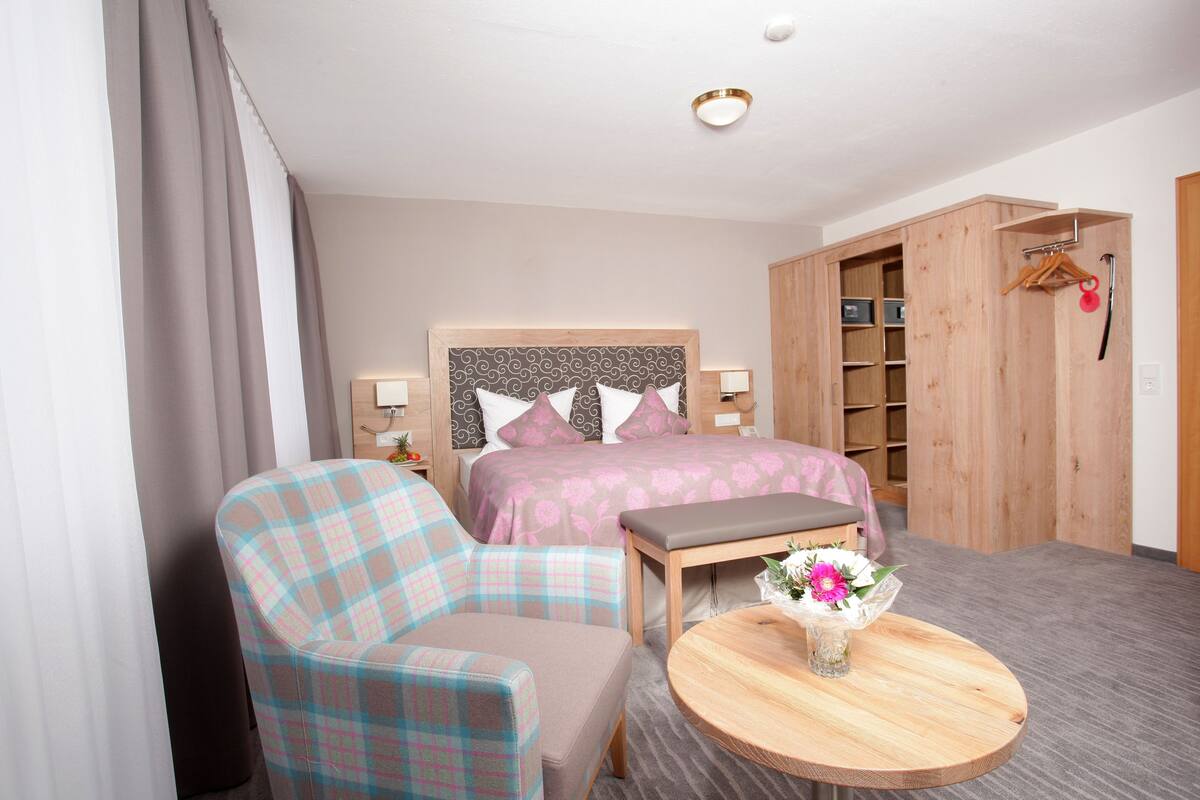 Quarto de hotel com uma cama, uma cadeira, uma mesinha com um vaso de flores e um guarda-roupa.