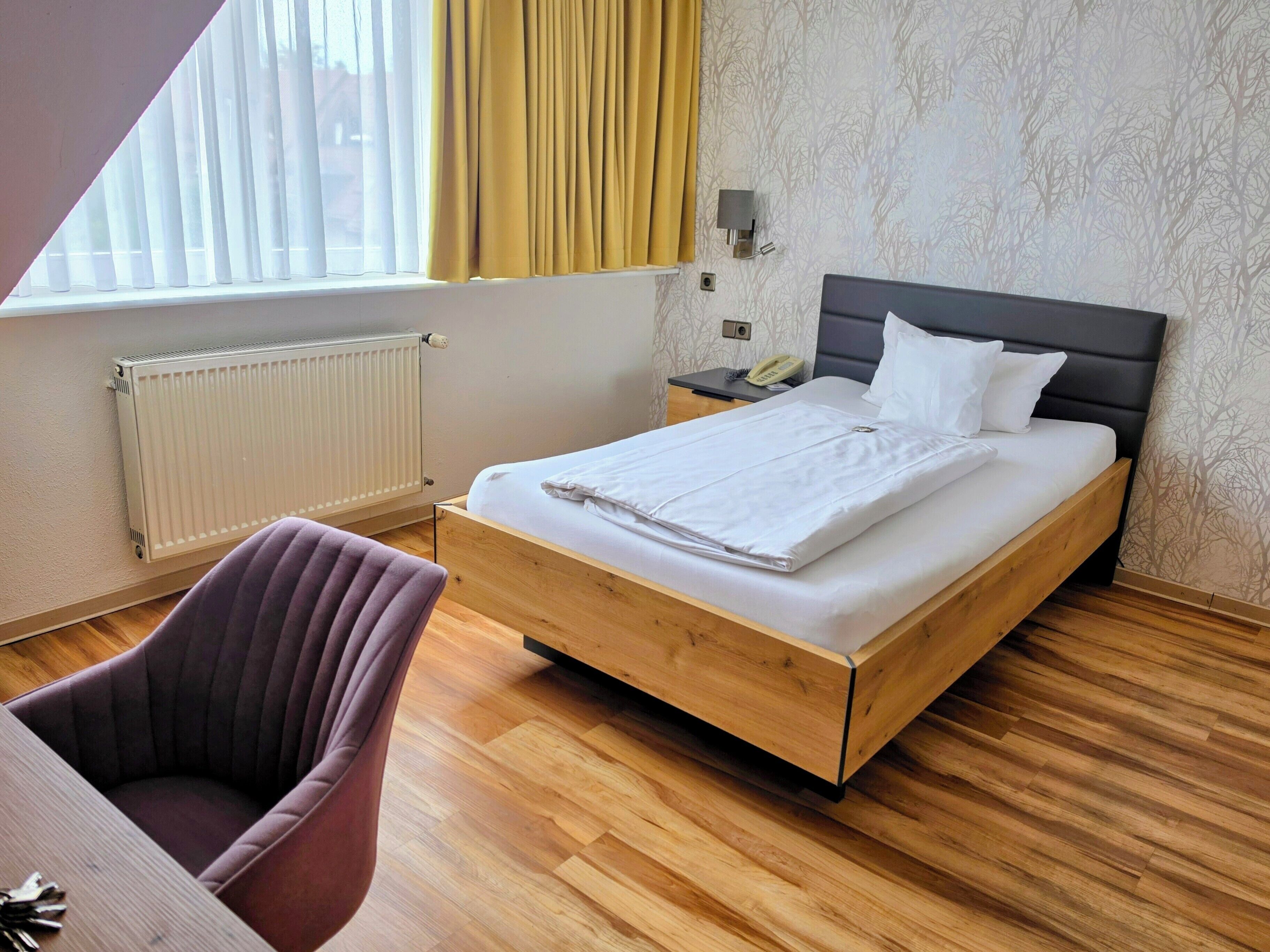 Een net opgemaakt bed met houten frame, een nachtkastje met lamp en een stoel in een kamer met houten vloer en een radiator.