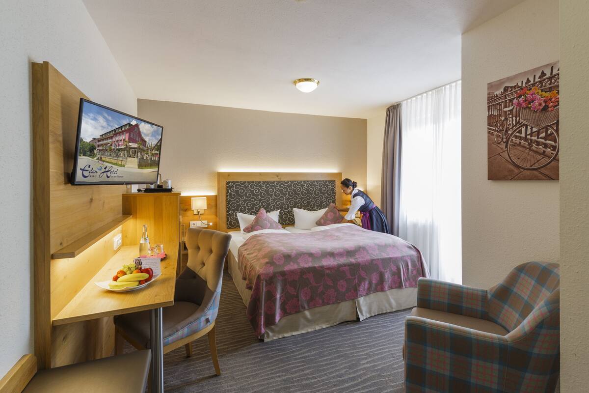 Quarto de hotel com uma cama, uma mesa com uma travessa de fruta, uma cadeira, uma televisão e um quadro na parede.