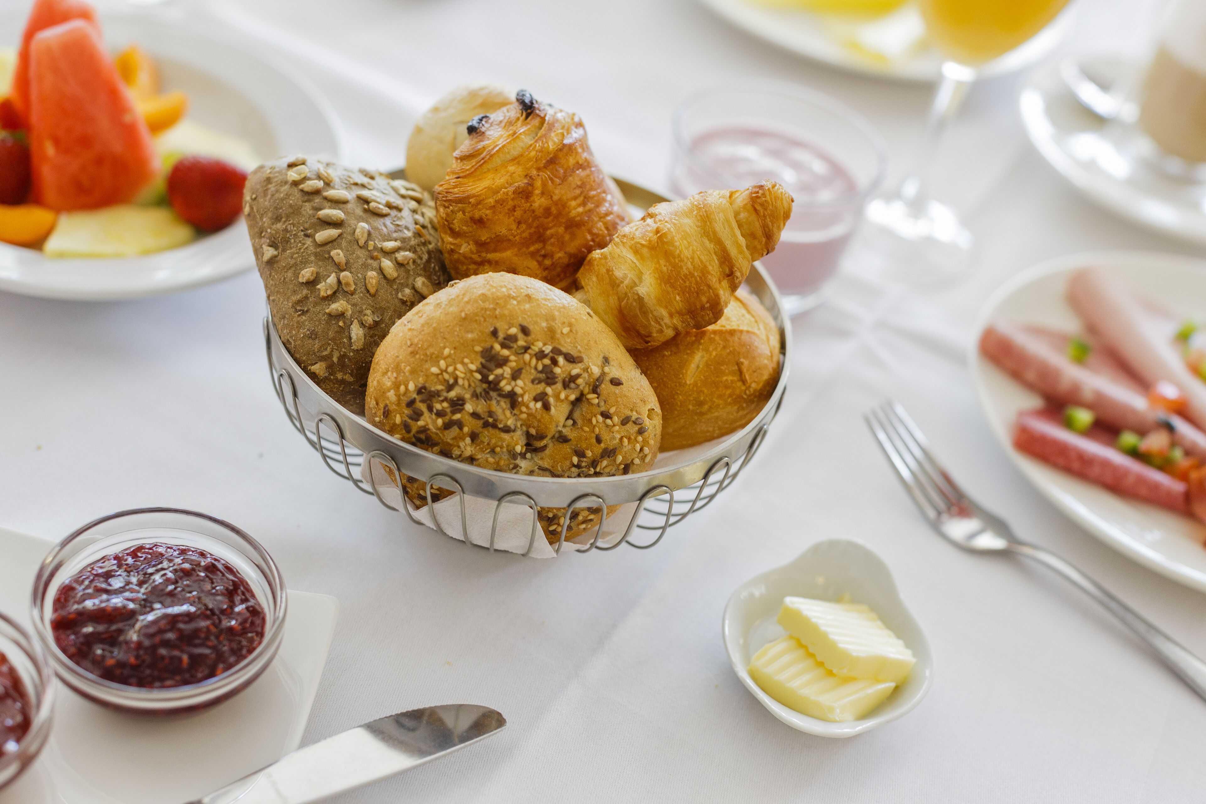 Une table mise pour le petit-déjeuner, avec un panier rempli de différentes sortes de pains et de viennoiseries, une assiette de saucisses et un petit bol de beurre.