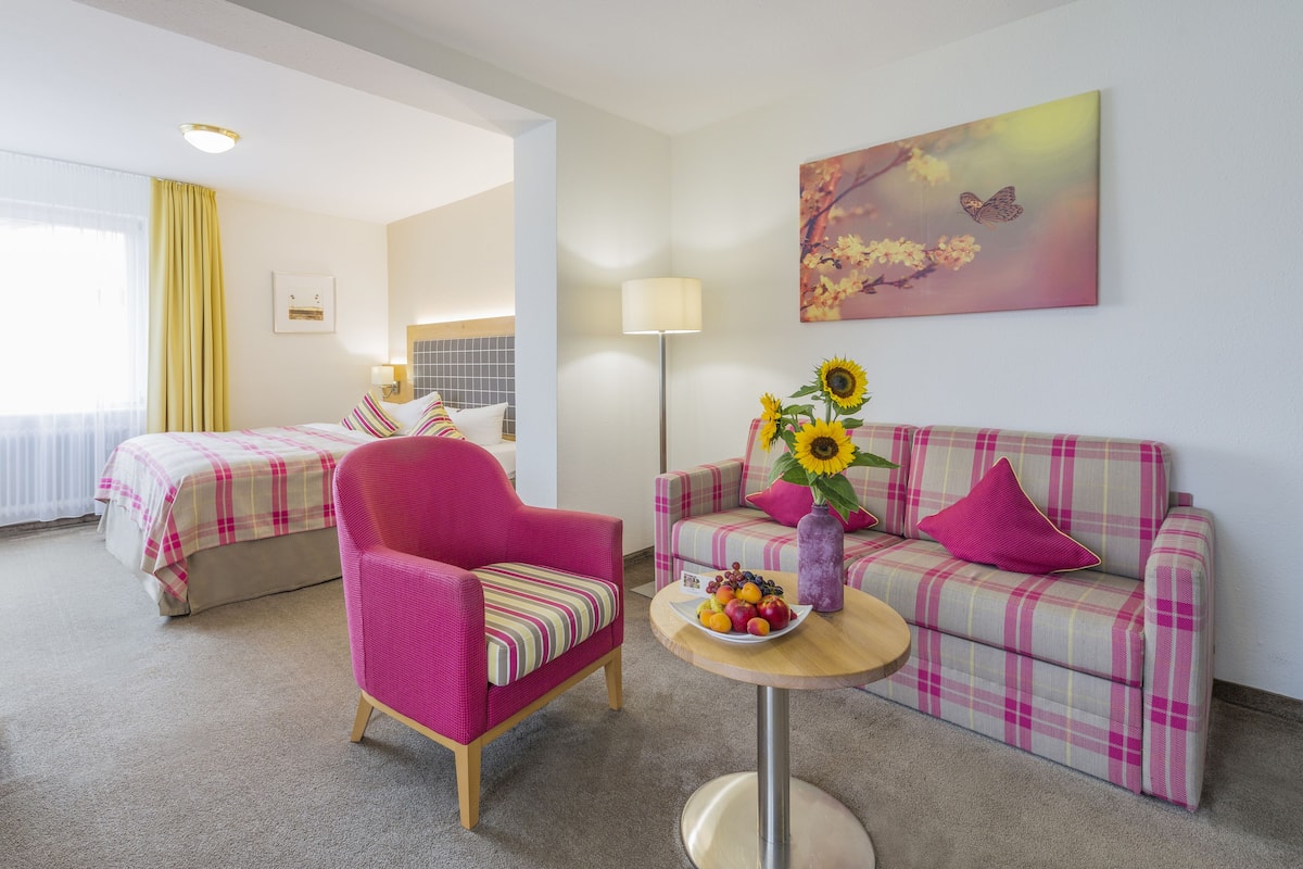 Quarto de hotel com cama, poltrona rosa, sofá xadrez, mesa redonda pequena com um vaso de girassóis e um quadro de borboleta.