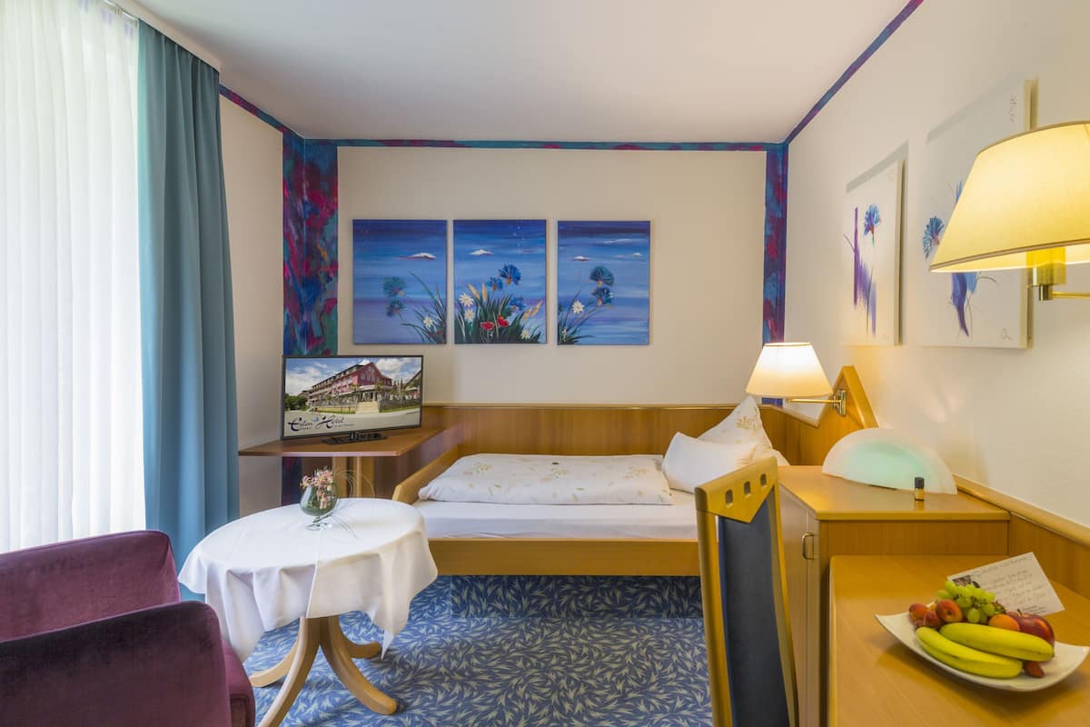 Quarto de hotel com cama, uma pequena mesa com um vaso, uma cadeira, escrivaninha com luminária e um prato de frutas.