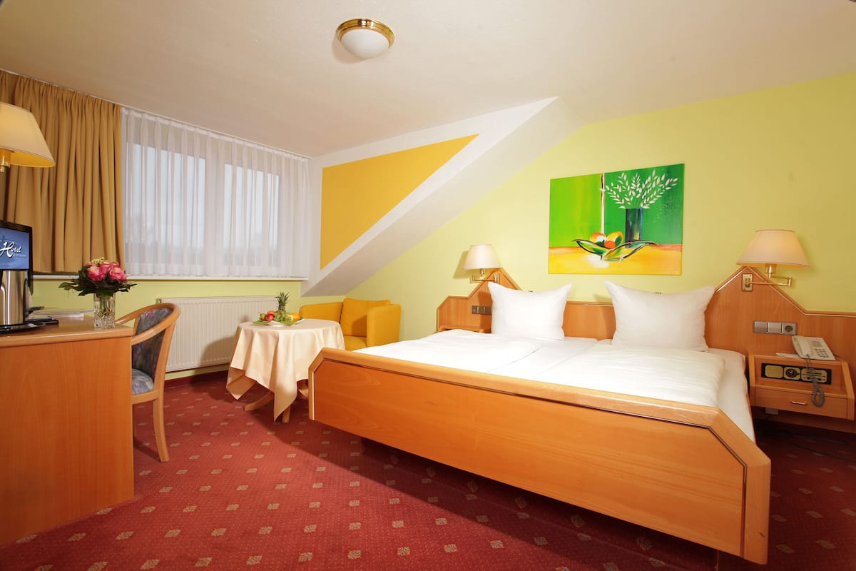 Quarto de hotel com uma cama grande, uma escrivaninha com um computador, uma mesinha com um vaso de flores e um quadro na parede.