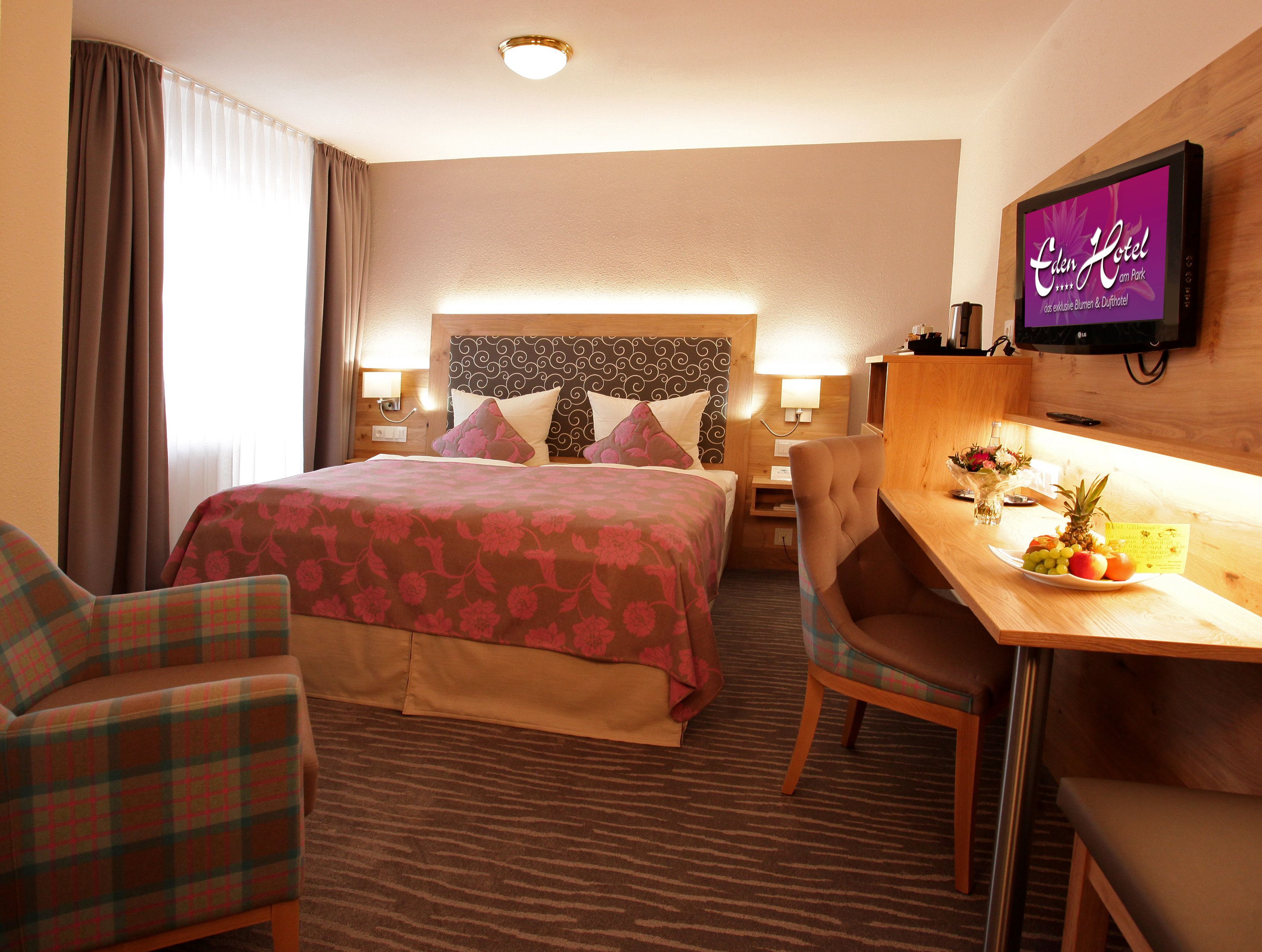 A hotel room with a bed, two chairs, a desk with a fruit plate, a TV, and a headboard with a patterned design.
