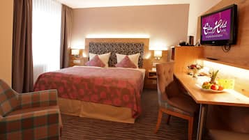 A hotel room with a bed, two chairs, a desk with a fruit plate, a TV, and a headboard with a patterned design.