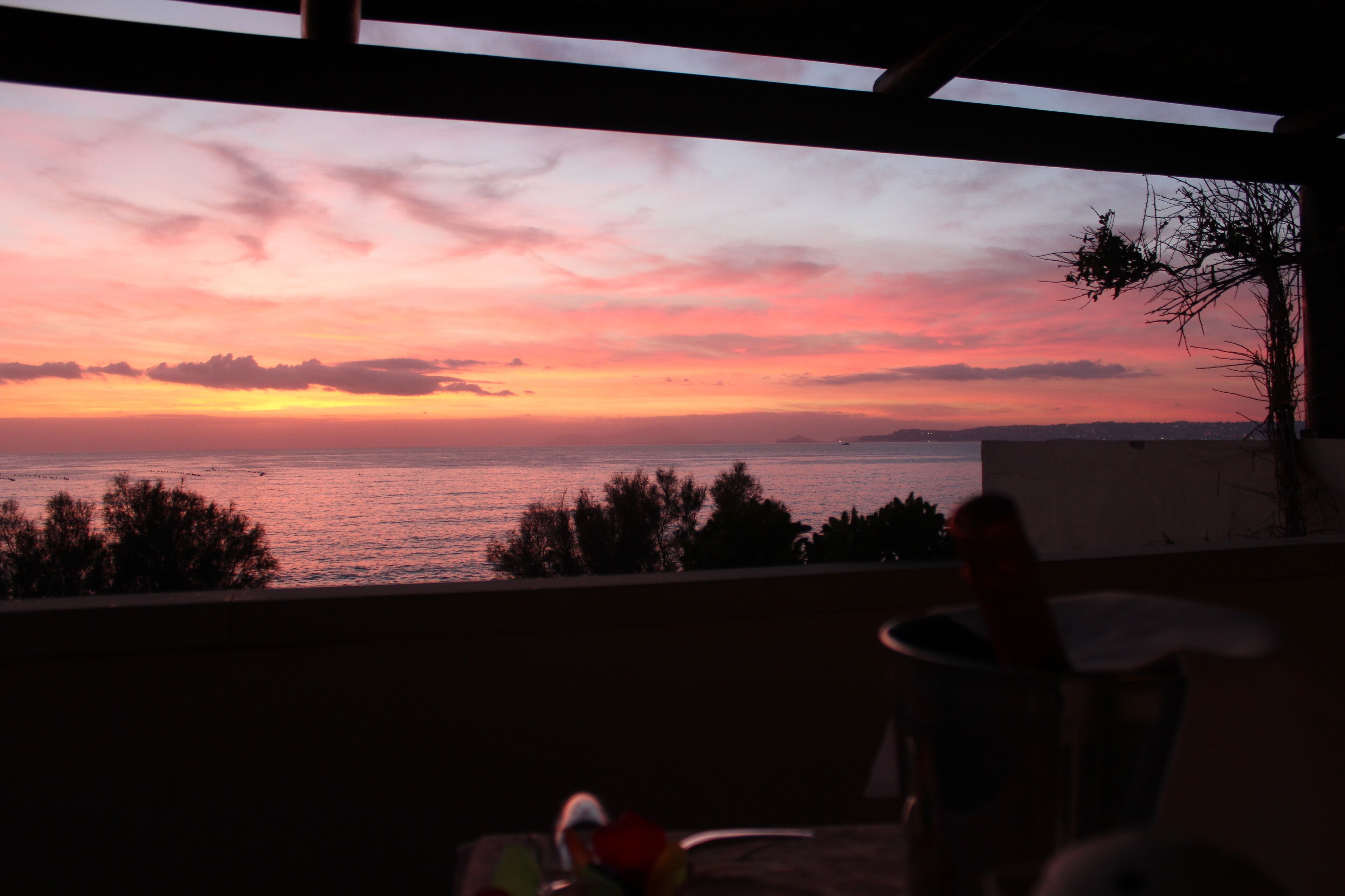 A sunset view from a balcony overlooking the sea, with a chair and table in the foreground.