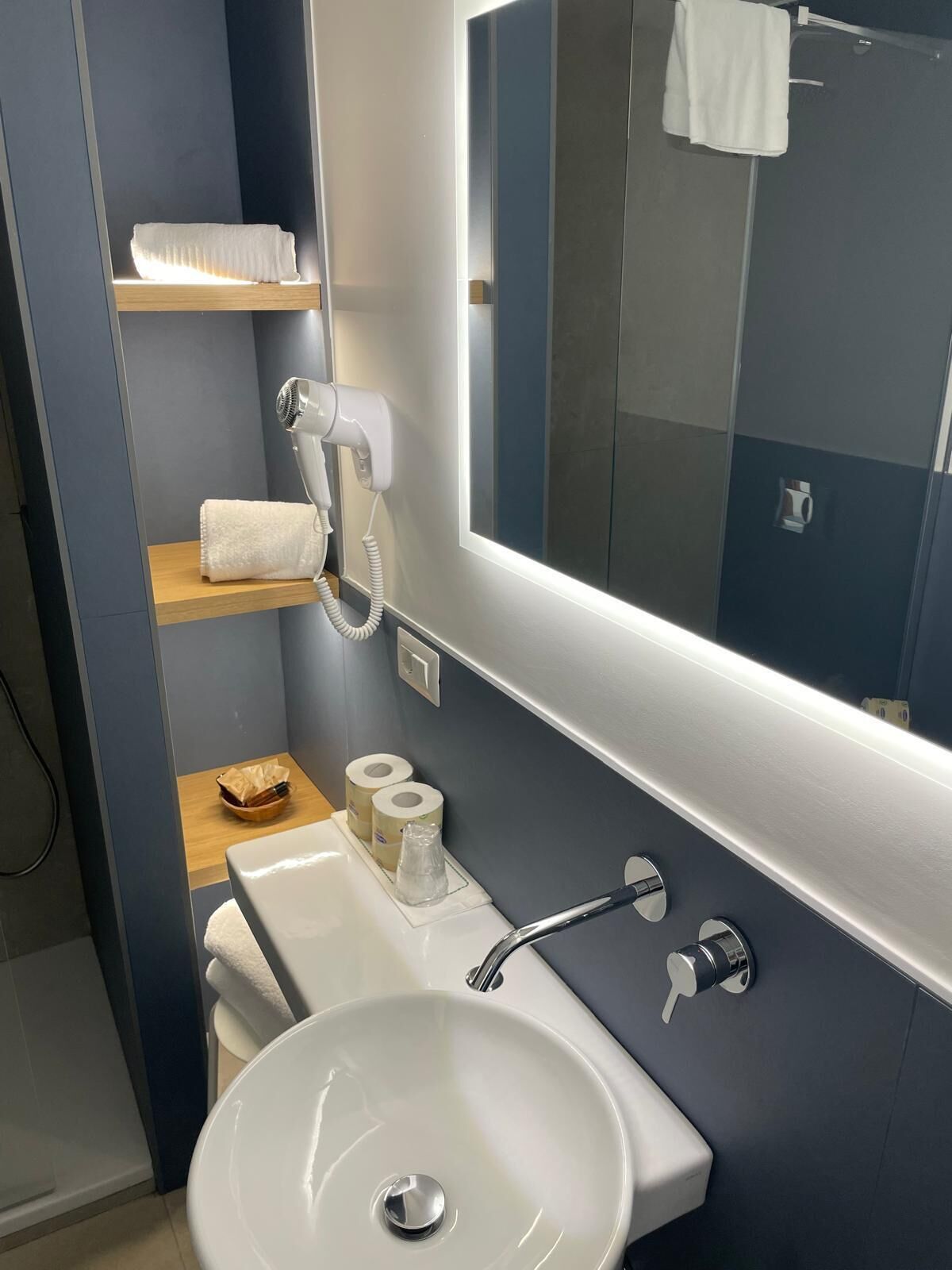 Un baño moderno con un lavamanos blanco, un espejo, un perchero para toallas y un estante con artículos de aseo.