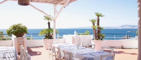 Un área de comedor al aire libre con mesas y sillas, techo de paja y vista al mar.