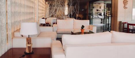A modern living room with white sofas, a wooden side table, and a lamp. There is a glass partition with a plant and a coffee machine.