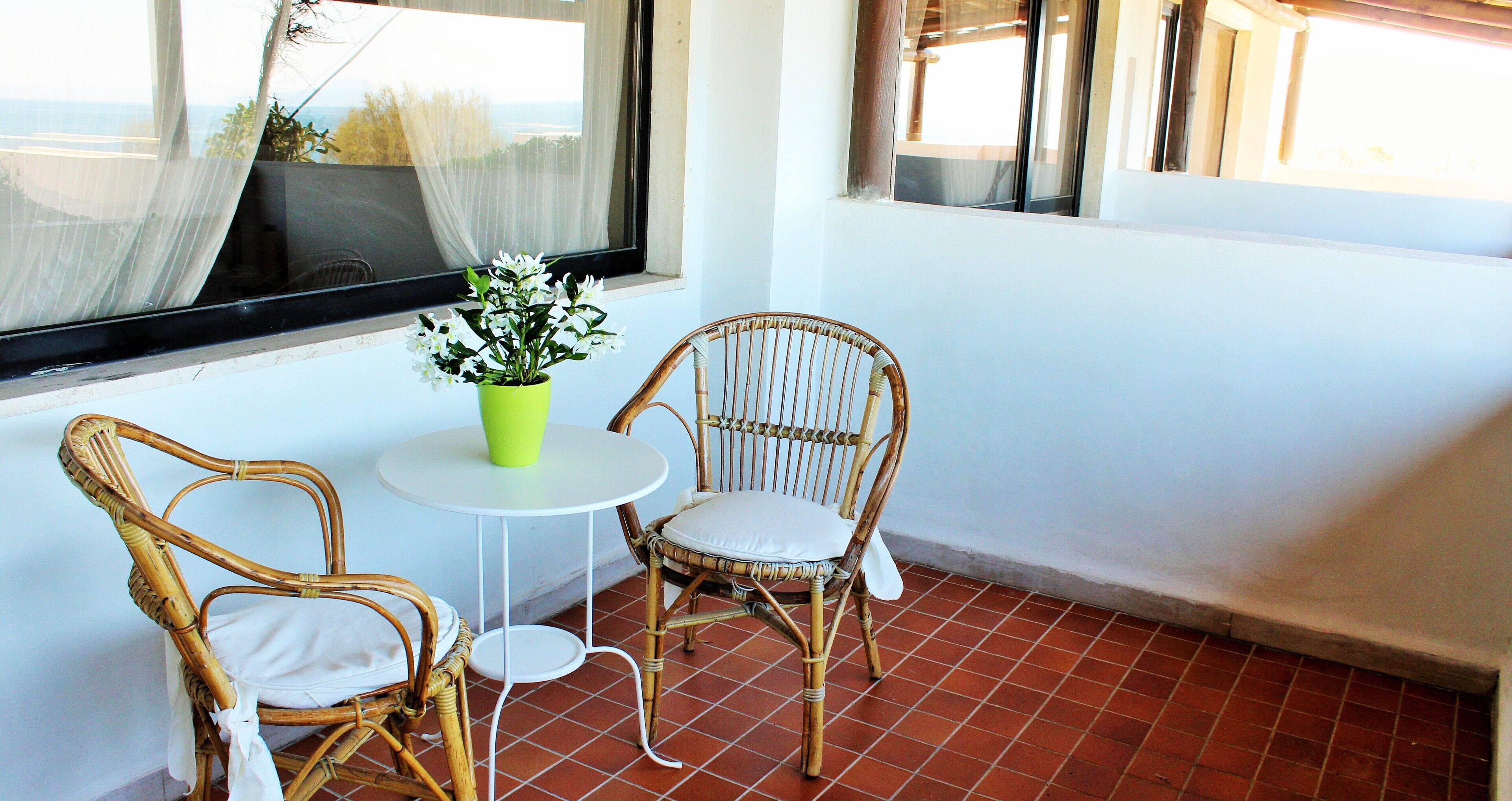 A balcony with two wicker chairs and a small round table, a potted plant, and a view through a window.