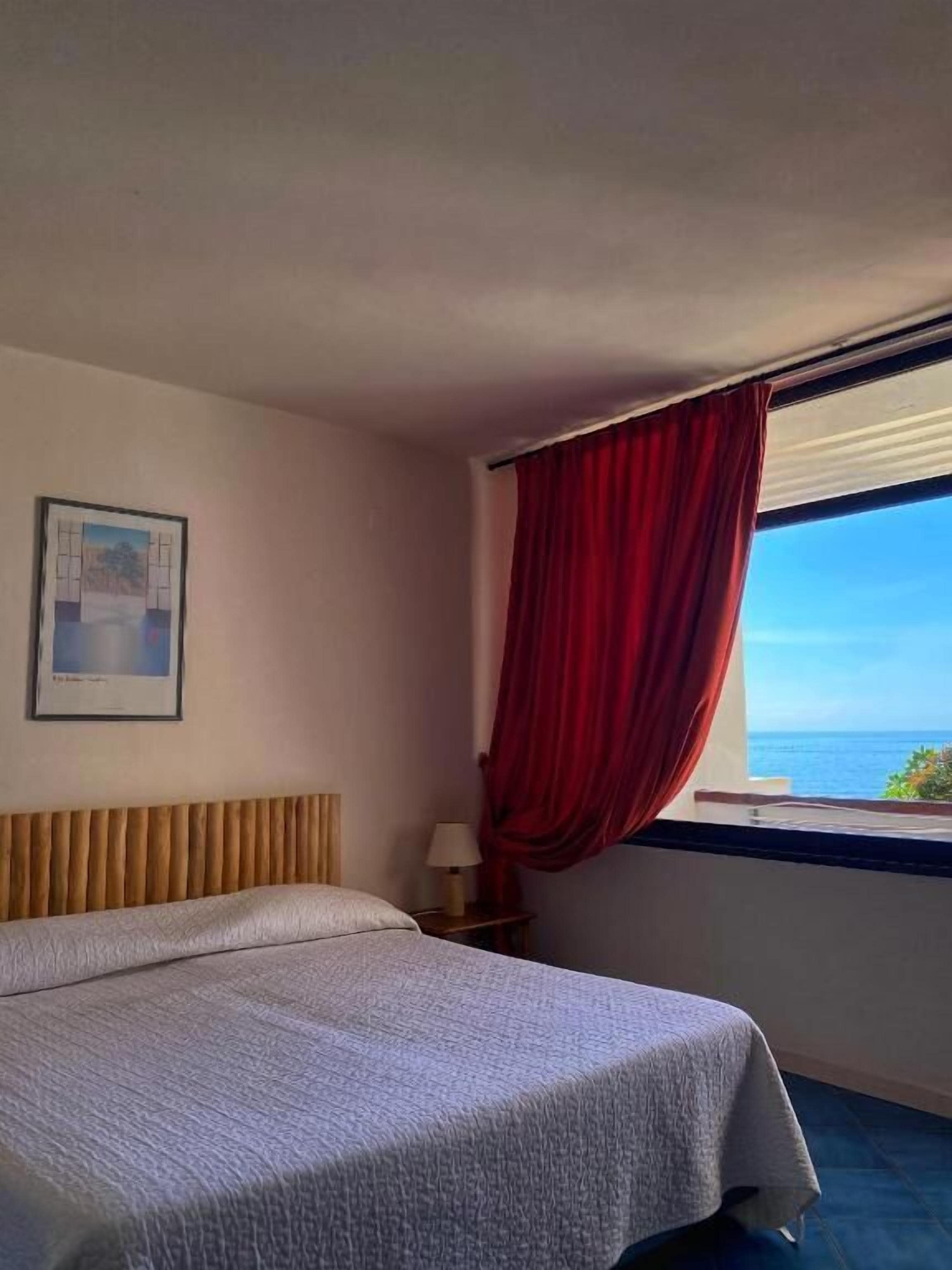 Ein Hotelzimmer mit Bett, Nachttisch, Lampe, einem gerahmten Bild an der Wand und einem Fenster mit rotem Vorhang.