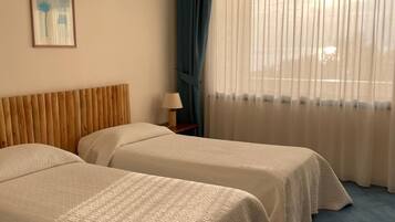 Ein Hotelzimmer mit zwei Betten, einem hölzernen Kopfteil, einem Nachttisch mit Lampe, einem gerahmten Bild an der Wand und einem großen Fenster mit durchsichtigen Vorhängen.
