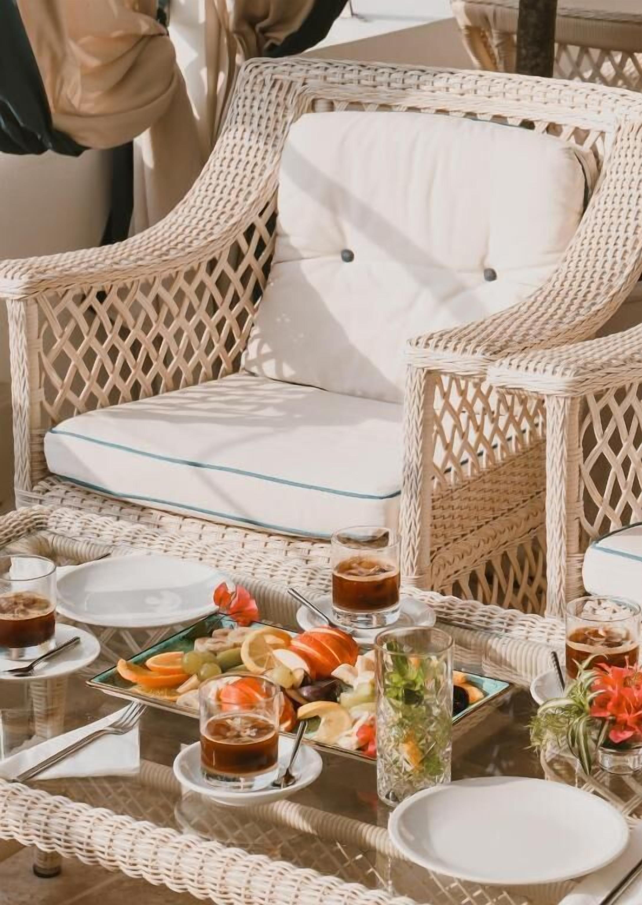 A wicker dining set with a glass table, plates, cutlery, and a variety of fruits and beverages.