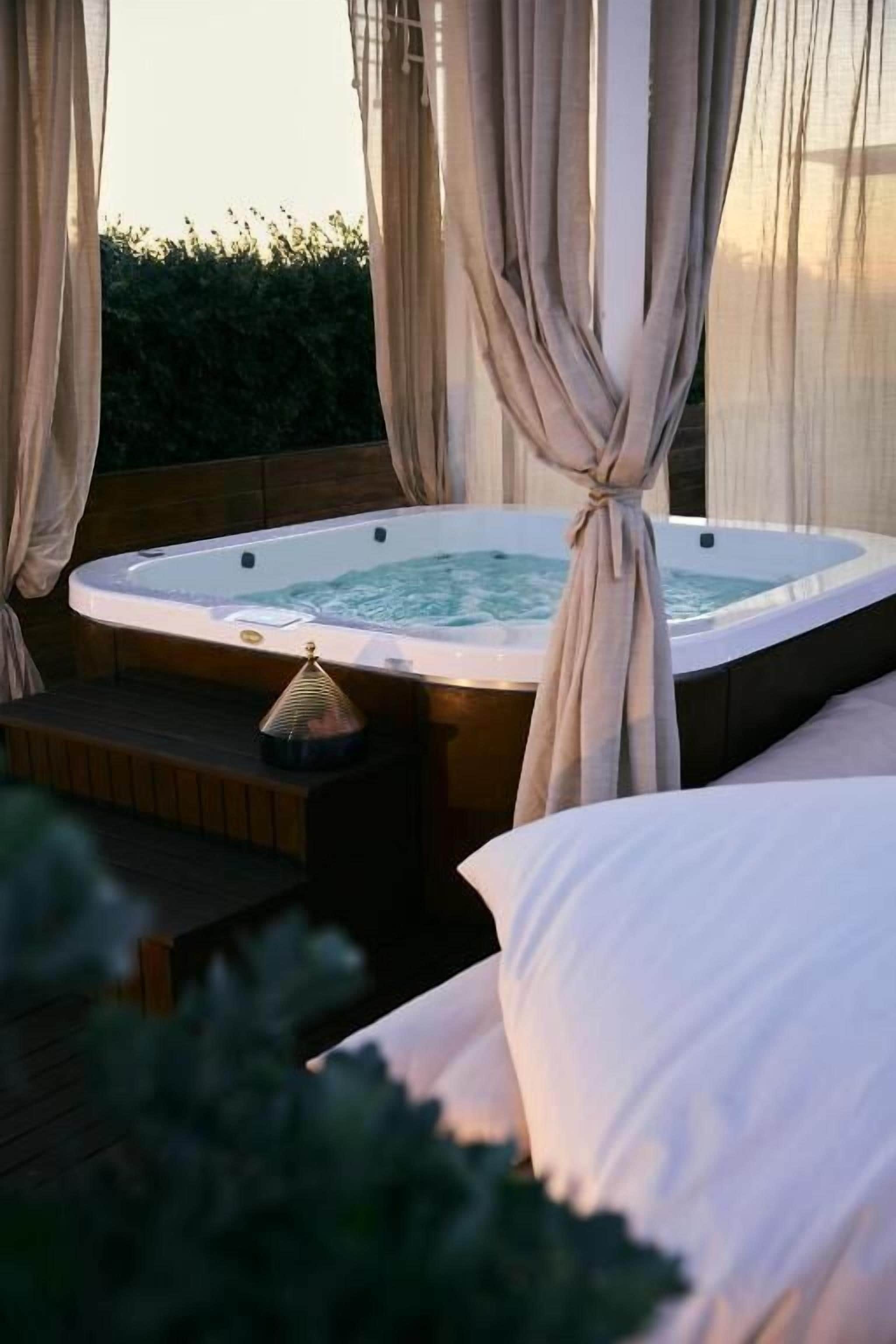 A hot tub with a wooden bench and a small table with a lamp, surrounded by curtains and a view of greenery.