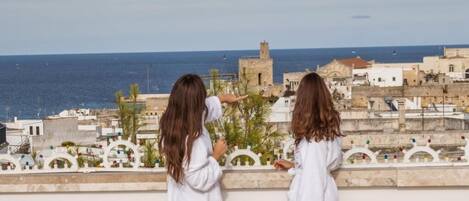 Two individuals on a balcony overlooking a coastal town with historic buildings and a view of the sea.