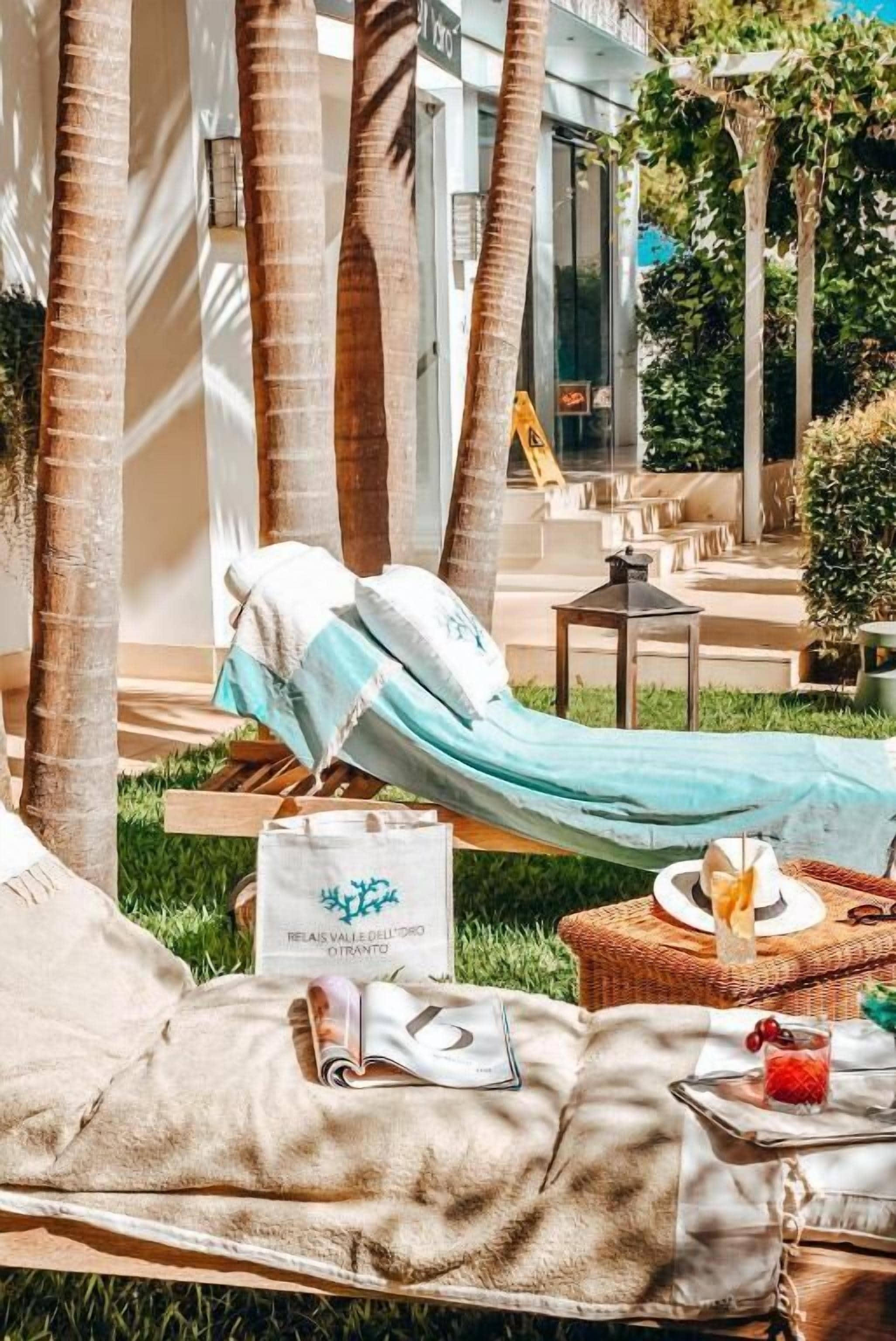 A hammock lounge area with a wicker table, a glass of drink, and a sign reading "RELAIS VALLE DELL'ORO OTRANTO".
