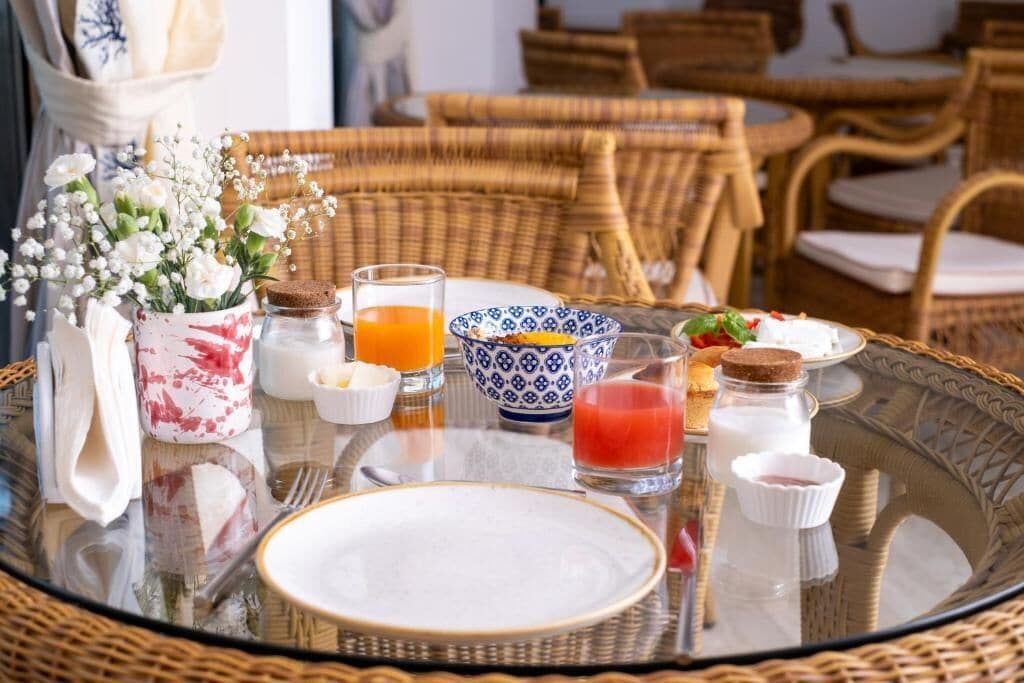 Un desayuno con mobiliario de mimbre, una mesa de vidrio y diferentes productos para el desayuno, incluyendo un jarrón con flores, un vaso de jugo de naranja y un tazón de cereales.