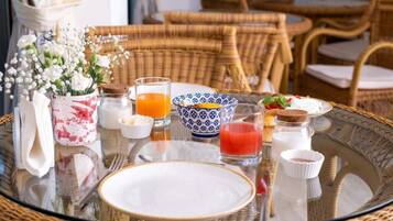 Un desayuno con mobiliario de mimbre, una mesa de vidrio y diferentes productos para el desayuno, incluyendo un jarrón con flores, un vaso de jugo de naranja y un tazón de cereales.