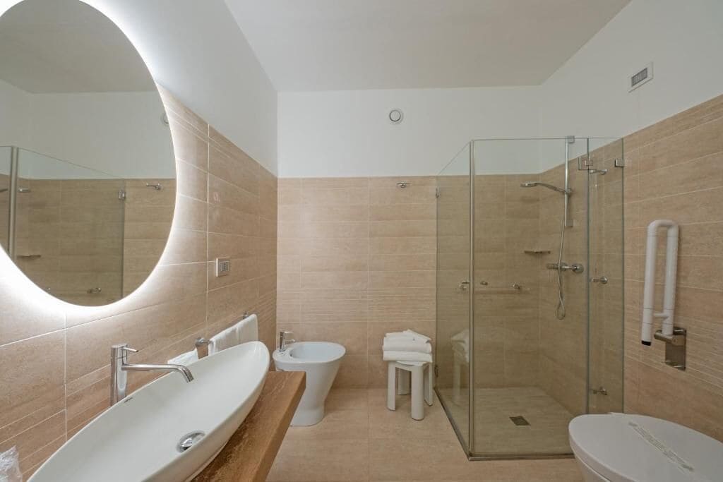 A modern bathroom with a large oval mirror, a freestanding sink, a bidet, a toilet, and a glass-enclosed shower.
