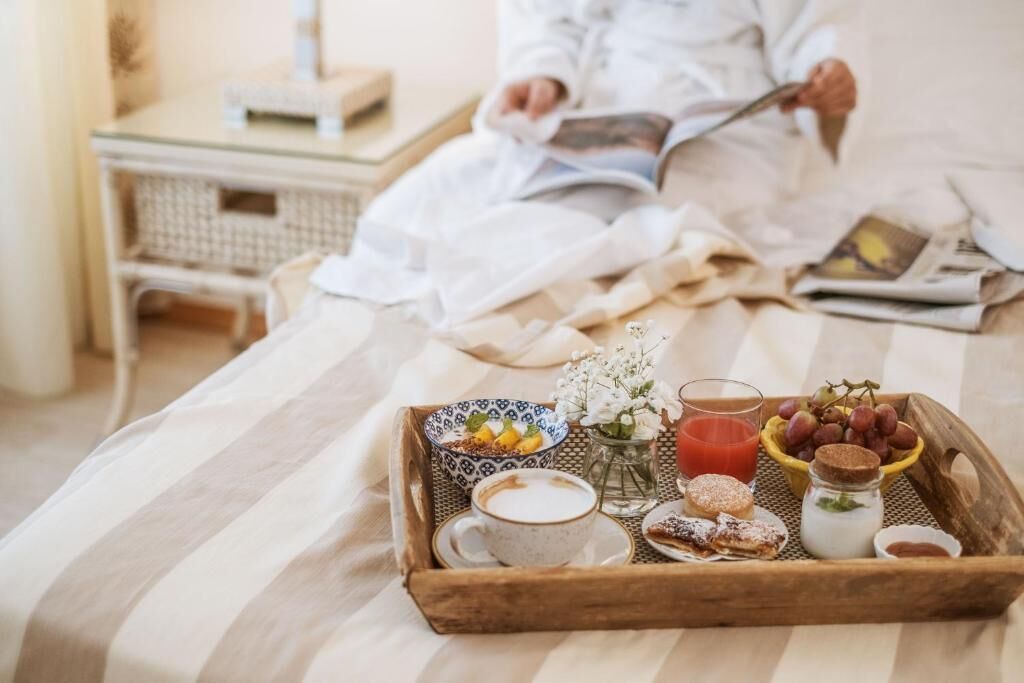 Bandeja de desayuno con café, pasteles y fruta sobre una cama, con un libro y una lámpara al fondo.