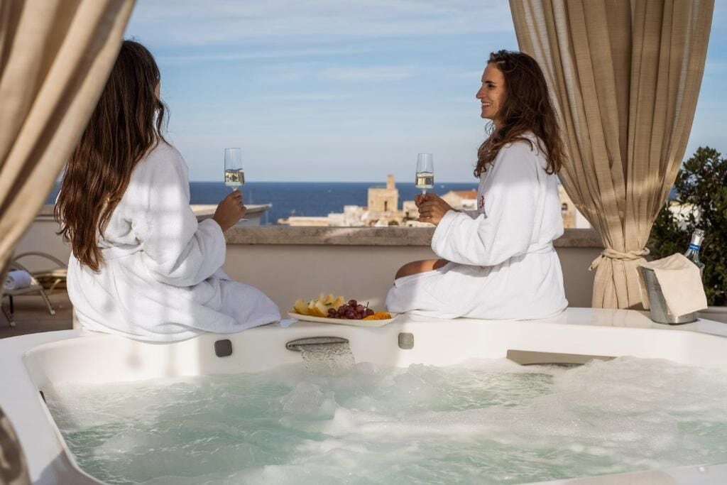 Zwei Personen in weißen Roben entspannen in einer Whirlwanne mit Blick auf das Meer und Gebäude im Hintergrund.