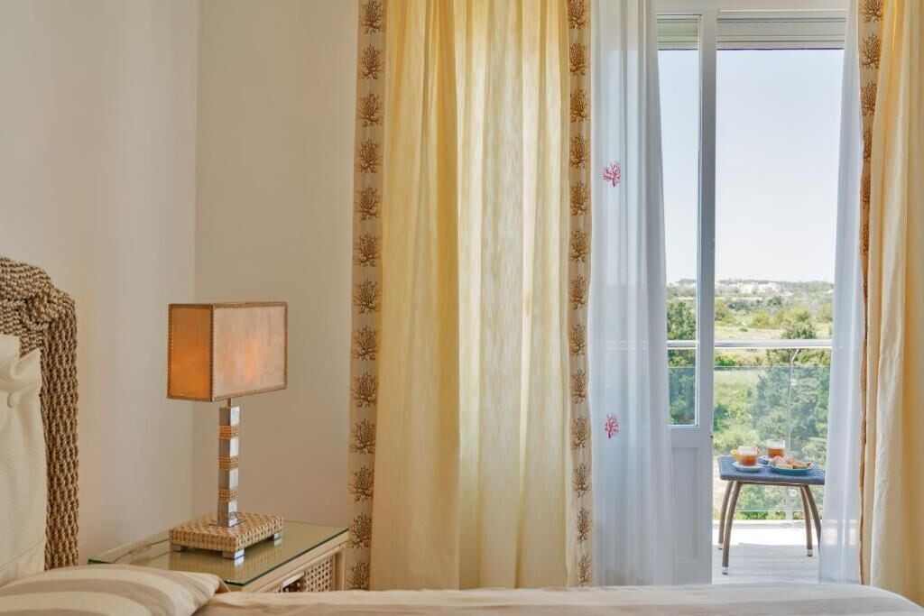 Habitación de hotel con cama, mesita de noche con lámpara, ventana con cortinas y balcón con mesa y comida.